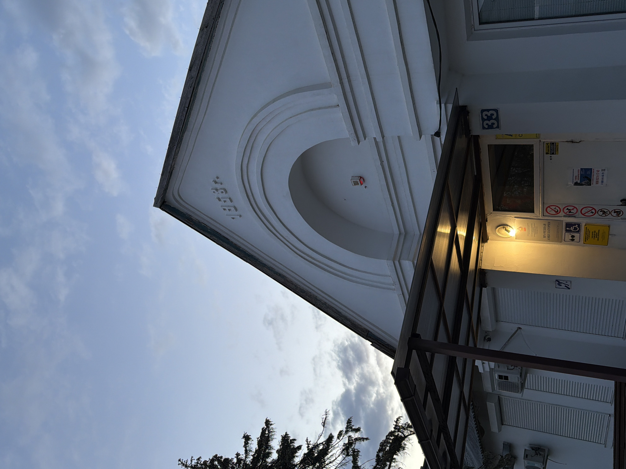 The image, taken from a low angle and rotated clockwise, depicts the exterior of a white building against a cloudy sky. The building, likely located in Yuzhny, Russia, features a classical or neo-classical architectural style.
In the upper-right section of the frame (when re-oriented), a prominent decorative element of the building's facade is visible: a pediment-like structure with an arched recess and multiple horizontal relief bands. The year "1935" is carved into the flat surface of this architectural feature.
Below this, an entrance area is illuminated by a warm yellow light fixture mounted under a brown, metal-framed awning with translucent panels. Next to the door, the blue building number "33" is affixed. Several signs are visible on the wall adjacent to the entrance. One clearly reads "ВРАЧЕБНАЯ АМБУЛАТОРИЯ" (Medical Outpatient Clinic) in Cyrillic script. Other signs include a universal disabled access symbol and common prohibition signs (no smoking, no dogs, no littering). Partial phone numbers are also discernable.
Further down the facade, two outdoor air conditioning units are mounted, and windows are covered by rolled-down security shutters. The sky, visible in the upper-left of the original image, is overcast with scattered clouds, indicating a time of day such as early morning or evening, when artificial illumination is necessary. A dark, coniferous tree is partially visible at the very bottom of the frame. No people or specific activities are present in the static scene.