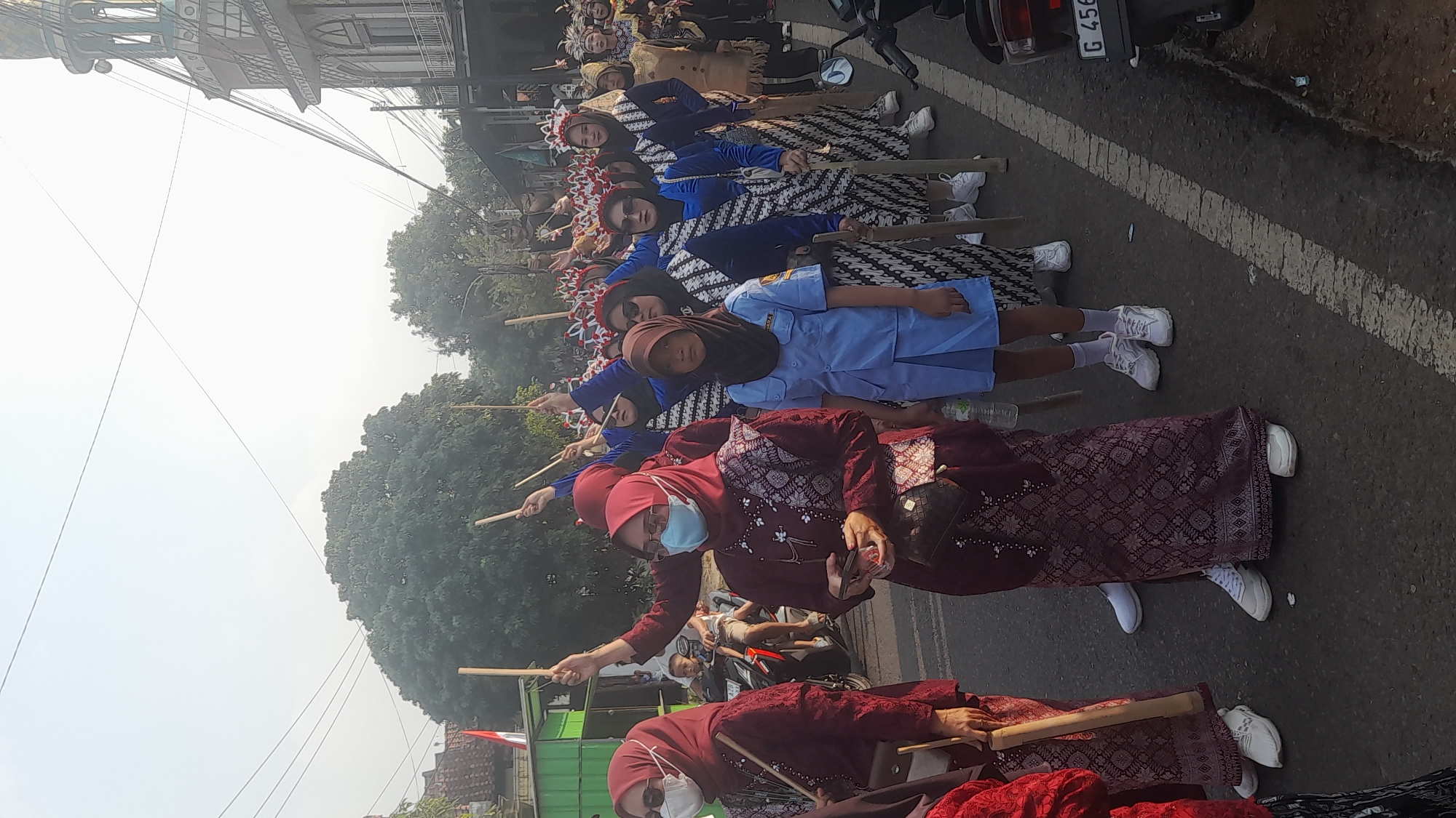 A procession is taking place on a street in Lemahabang, Indonesia. The foreground features a group of people, predominantly women, dressed in traditional attire. The women on the left wear maroon outfits with patterned skirts and headscarves, some wearing face masks. They are holding wooden staffs. To their right, a young girl in a light blue school uniform walks forward, also holding a wooden staff. Behind them, a line of women in blue and white patterned outfits and headscarves are also participating, some carrying staffs. In the background, a mosque with an ornate minaret is visible on the right, and trees and buildings line the street on the left. The sky is bright and hazy, suggesting it is daytime. A motorcycle is parked on the right side of the road.