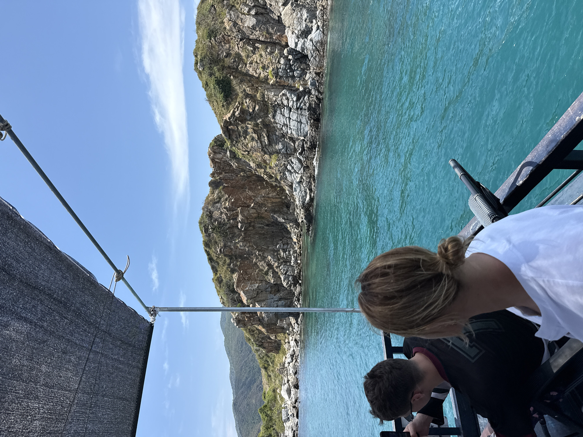 The image shows a partial view of a boat near a rocky coastline, with a view towards land. Two people are visible on the boat's edge; a woman with blonde hair pulled into a bun is in the foreground, wearing a white shirt. A younger person, possibly male, is next to her, wearing a black shirt with maroon trim. They appear to be looking at something. The water is turquoise. The coastline features cliffs with visible rock striations and some green vegetation. A secondary, lower hill is visible behind the main cliff. A gray sunshade is visible in the foreground, attached to a metal frame. The location is Bac Nha Trang Ward, Vietnam, based on geographical similarities.