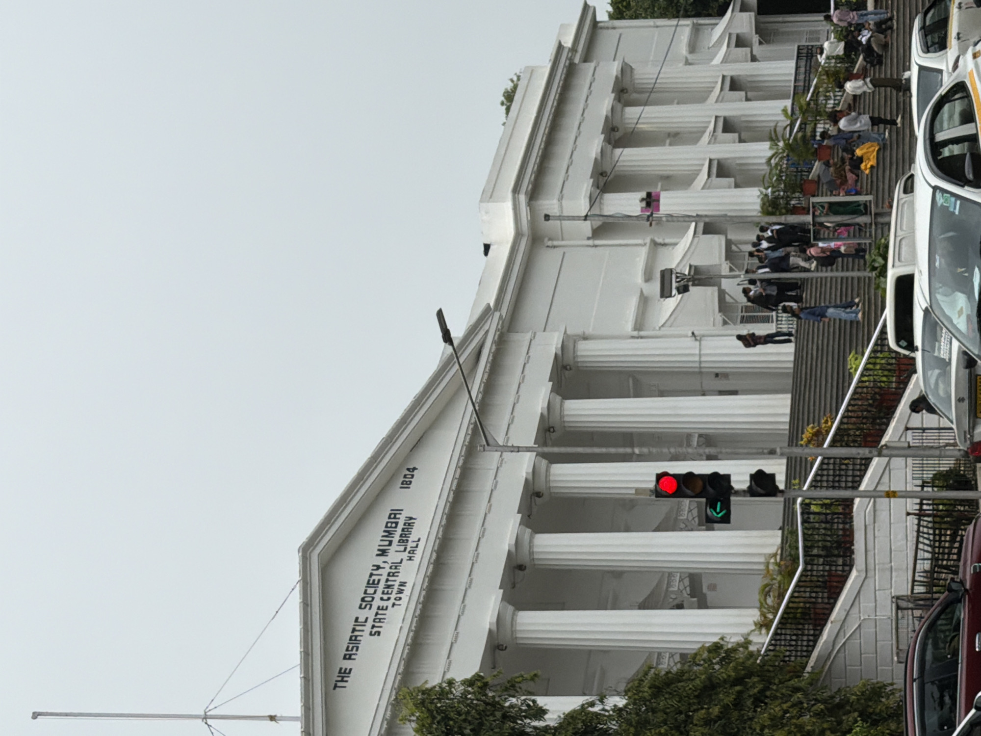 The image captures the front facade of "The Asiatic Society, Mumbai, State Central Library, Town Hall," a prominent building in Mumbai, India. The architecture features a grand entrance with a series of large, fluted white columns supporting a pediment with inscriptions. "THE ASIATIC SOCIETY, MUMBAI," "STATE CENTRAL LIBRARY," "TOWN HALL," and "1804" are clearly visible on the building's facade.
A set of wide, white stone stairs leads up to the library entrance. Several potted plants are strategically placed along the railings of the stairs. A traffic light is positioned in the foreground, showing a red light illuminated and a green arrow pointing left. Cars are visible on the road in front of the building, including a red car to the left and white cars to the right.
A group of people are gathered on the steps, some standing and some sitting. A few individuals are also seen on the sidewalk in front of the stairs. The sky is overcast, suggesting a cloudy day. Lush green trees are visible to the left and right edges of the frame, adding a touch of nature to the urban landscape. The overall scene depicts a busy public space in front of a historic institution in Mumbai.