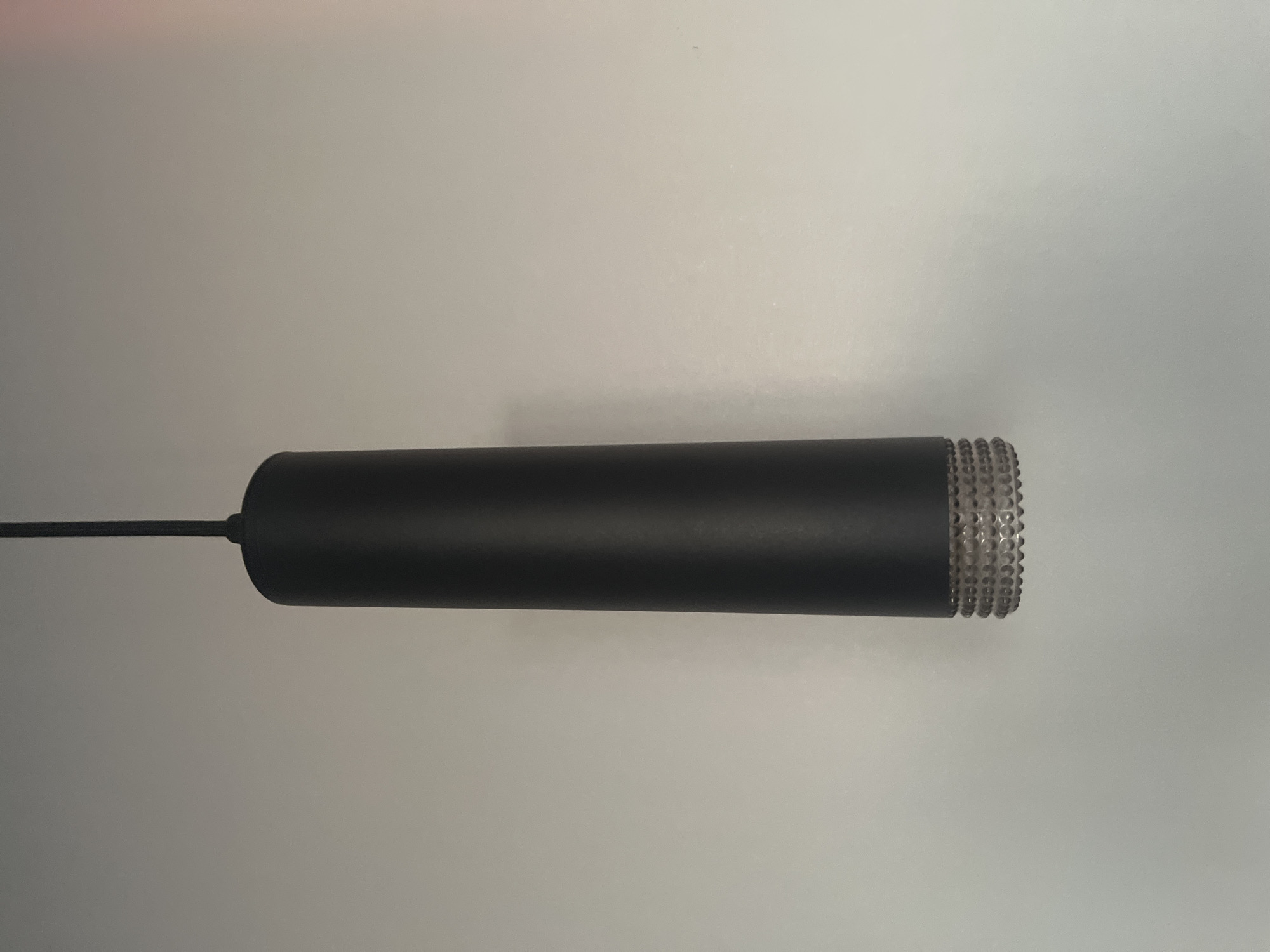The image shows a black cylindrical object with a textured, clear end. The object is suspended by a thin black cord against a plain, light gray background. The lighting suggests an indoor setting with diffuse, overhead illumination. There are no people or other objects visible in the frame, and no discernible text. The focus is entirely on the suspended object.