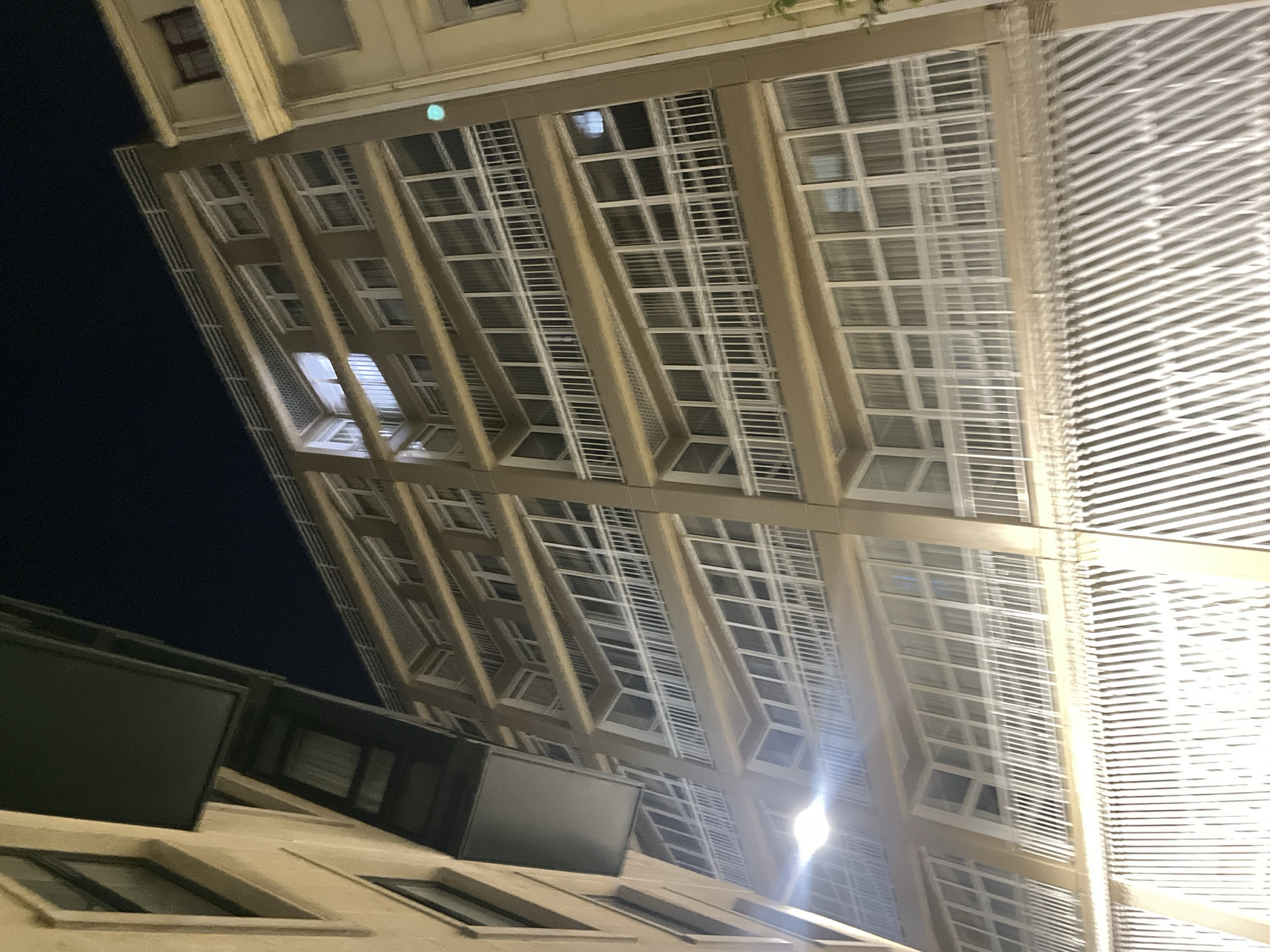 The image shows the architecture of a building in A Coruña, Spain, specifically looking upwards at a structure with a grid-like façade. The grid is created by repeating rectangular windows and parallel white horizontal lines, which seem to be Venetian blinds. The supports of these structures are painted beige. There is a second building to the side with windows that are visible on the bottom-left. The sky is visible in the top-left of the frame and is dark, implying it is night-time.