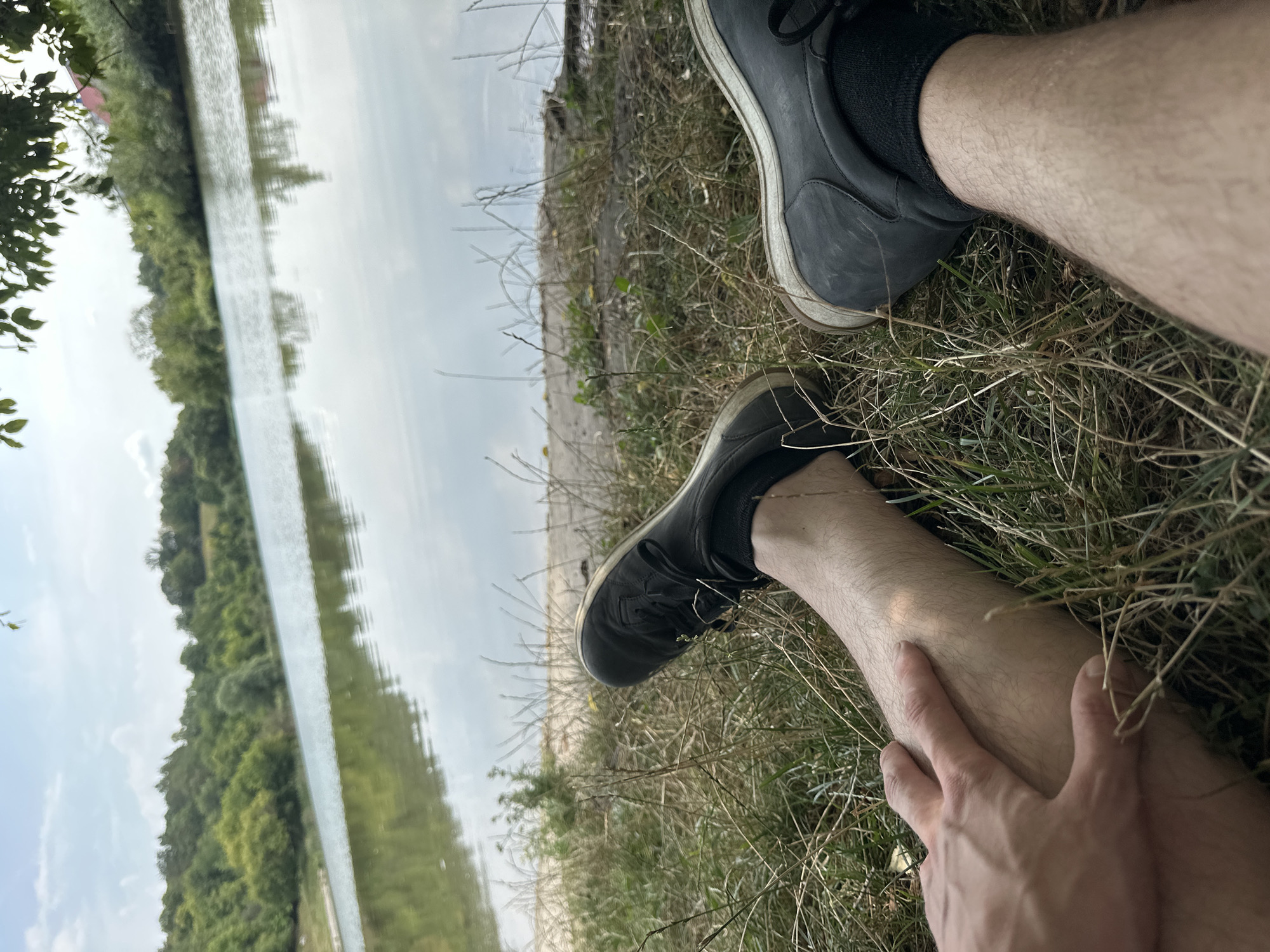 A first-person perspective shot shows a pair of legs wearing black shoes and socks, resting on dry grass and dirt next to a body of water. The water is calm and reflects the sky and the green, tree-lined hills in the background. The sky is mostly clear with some scattered clouds. The overall setting appears to be a natural, outdoor environment, likely on the outskirts of a town or village given the presence of trees and water. There are no visible buildings directly in the foreground, but hints of structures may be present in the distant treeline. The image captures a moment of relaxation or rest by the water.