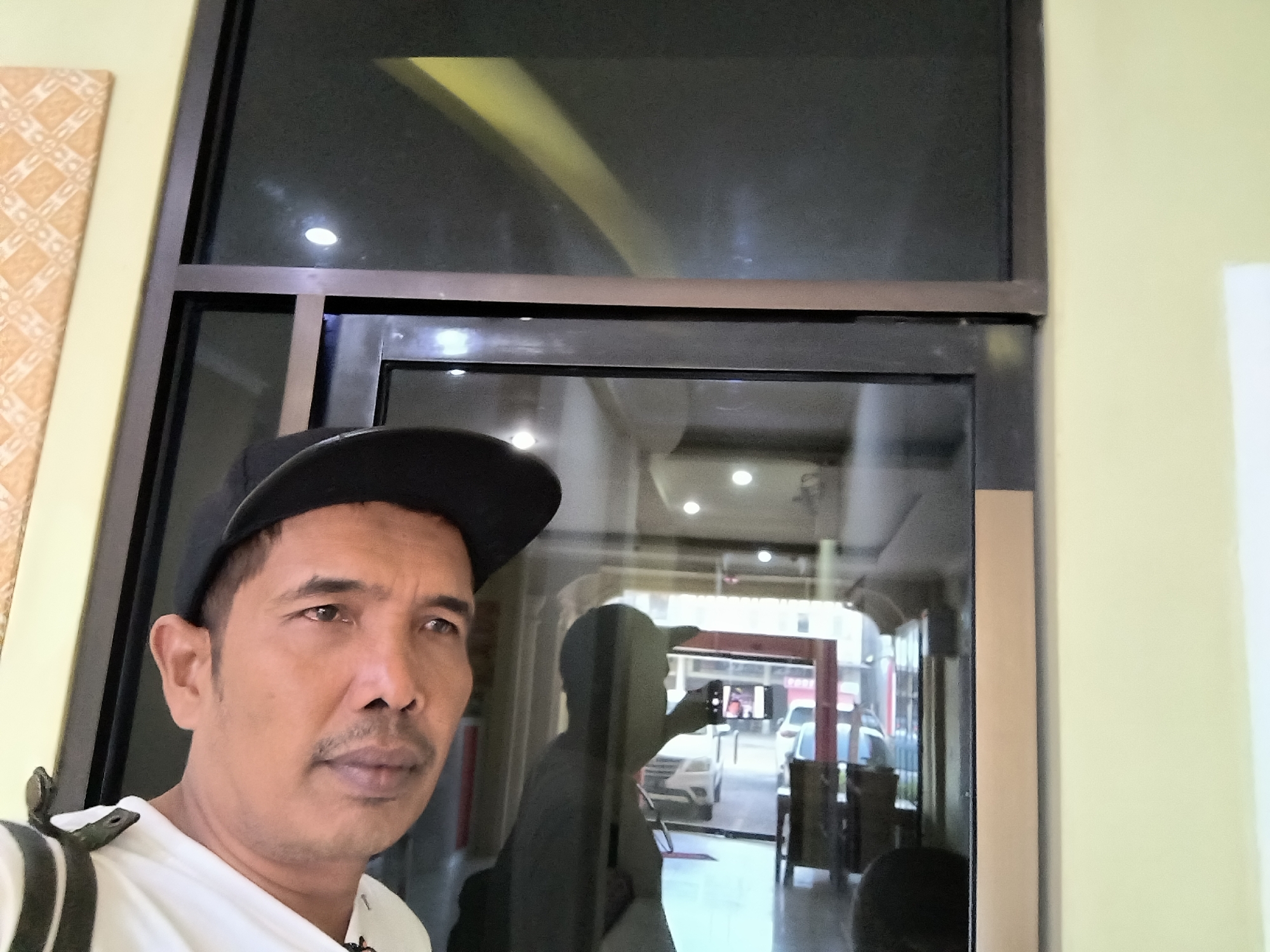 A man wearing a black baseball cap and a white shirt is taking a selfie in front of a reflective glass door. His face is partially visible in the foreground on the left side of the image. Through the glass, a reflection shows the man holding a phone to take the picture, and the interior of a building which appears to be a store or office with furniture and cars visible outside. The wall to the left of the man is light yellow with a patterned wallpaper. The scene takes place in an indoor setting with artificial lighting visible in the reflection. The overall impression is a candid moment captured by the subject himself.