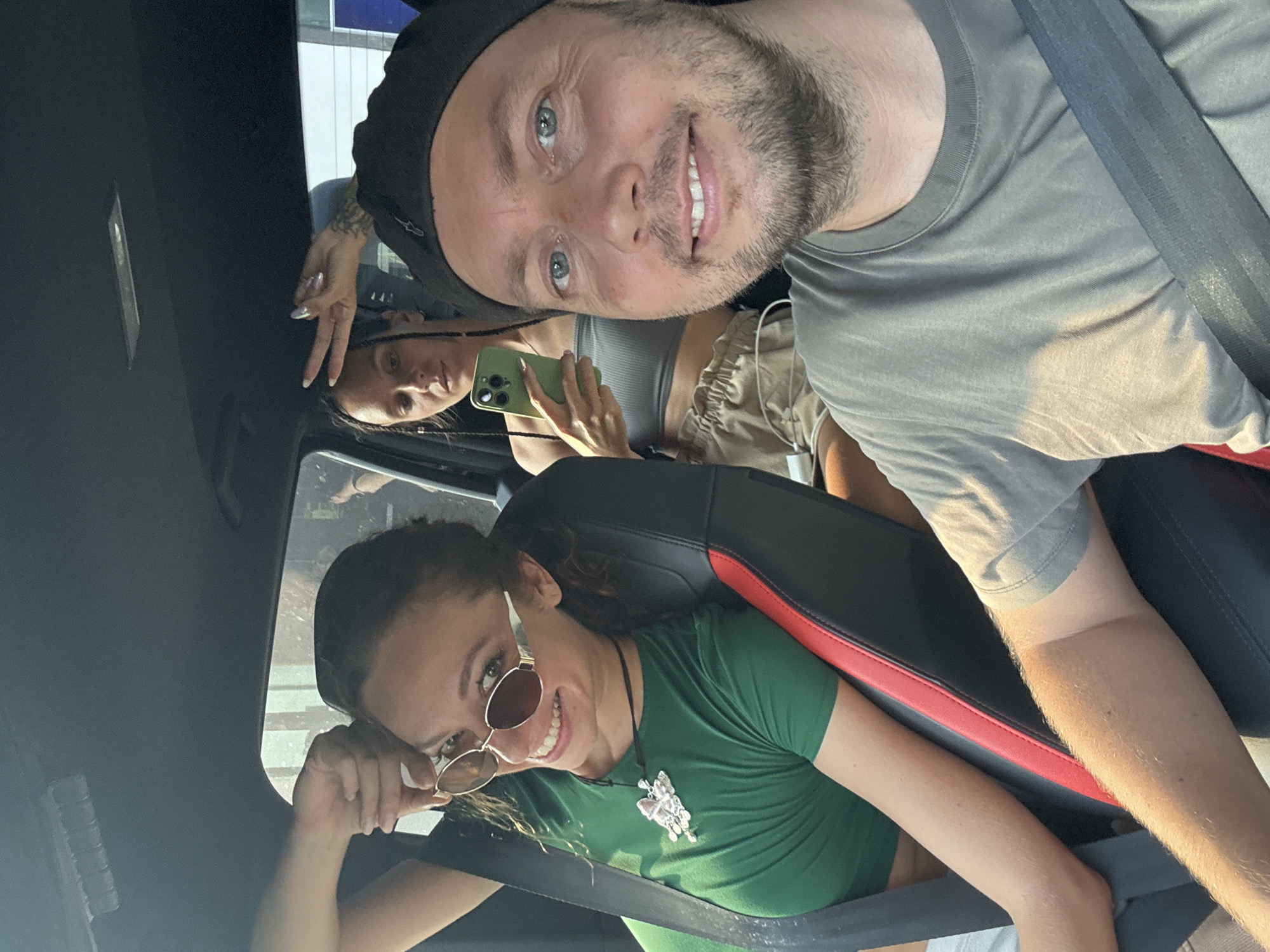 A rotated, bright selfie captures three smiling individuals inside a vehicle, likely a car. In the foreground, a man with a beard and blue eyes, wearing a black beanie and a grey t-shirt, smiles directly at the camera. He is secured by a seatbelt.
Behind him, a woman with braided dark hair, wearing a grey crop top and light-colored shorts, holds a green smartphone. Tattoos are visible on her left arm, and she is looking upwards, possibly at the man or the camera.
In the background, a third woman with curly dark hair and round, tinted sunglasses smiles broadly. She is wearing a green t-shirt and a prominent butterfly-shaped necklace, also secured by a seatbelt.
The scene is illuminated by bright sunlight, casting shadows on the subjects and the car's interior, indicating daytime. The interior features black and red upholstery on the car seats and a dark headliner. All three individuals appear cheerful and relaxed, suggesting a casual, happy moment during travel. No text is visible within the image. The provided location context is Hà Giang 1 Ward, Vietnam.