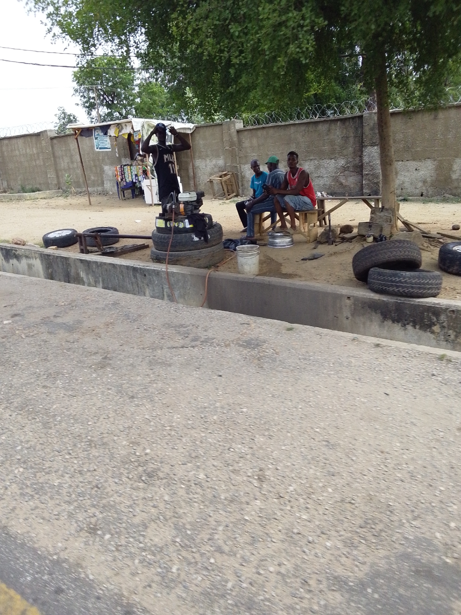 In Lokoja, Nigeria, an outdoor tire repair shop is positioned alongside a concrete road and elevated drainage channel. One man stands beside a tire repair machine constructed on a stack of tires. Three men are seated on a wooden bench under the shade of a large tree. A walled compound with barbed wire is visible in the background. A small kiosk selling items is adjacent to the tire shop. The scene is set during the daytime.