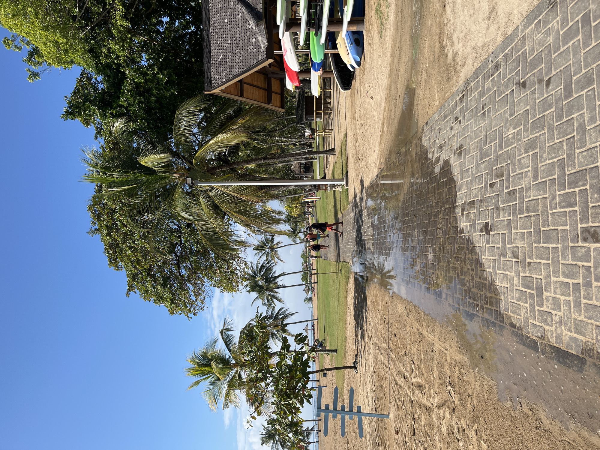 The image depicts a paved pathway on a sandy beach in Kuta Selatan, Indonesia, under a clear blue sky. The pathway, made of dark grey interlocking bricks laid in a herringbone pattern, is partially wet, reflecting the surrounding palm trees. To the left of the pathway is a sandy beach with footprints visible, and a blue directional signpost with multiple arms. Further left, lush green grass is visible, dotted with palm trees and beach umbrellas. Several people are walking along the pathway in the distance.
To the right of the pathway, a wooden structure with a dark tiled roof serves as a shelter, displaying an assortment of brightly colored surfboards. Beyond this, more palm trees and vegetation line the landscape, leading towards the ocean which is not clearly visible. The overall scene suggests a resort or recreational area on a tropical beach.