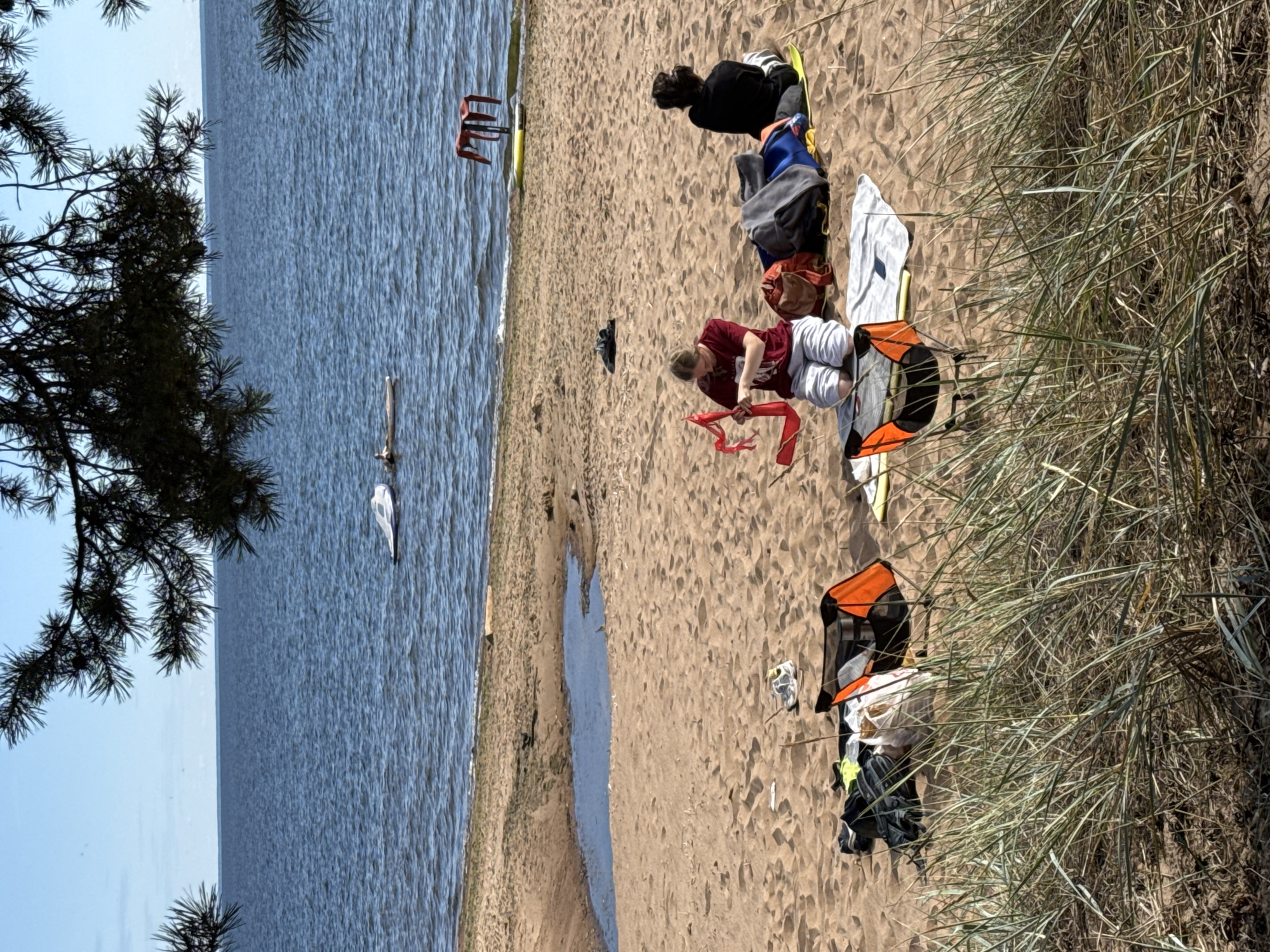 The image depicts a beach scene in Zelenaya Roshcha, Russia. In the foreground, tall grass partially obstructs the view of the sandy beach. Several items are scattered on the sand, including two foldable chairs, one orange and black, and another partially visible. A pile of bags and clothing lies near the orange chair.
Further back on the beach, two individuals are seated on the sand. One person, wearing a maroon t-shirt and white pants, is holding a red object, possibly a piece of fabric. Beside them is a collection of blankets and bags. The second individual, wearing a black hoodie and pants, is seated facing away from the camera.
In the background, the blue water of the sea stretches to the horizon under a clear sky. A small puddle of water is visible on the beach closer to the water's edge. Two white objects, possibly small boats or inflatable devices, are floating on the water, and a red object, appearing to be a chair or table, is situated on a yellow object on the beach near the water. The overall impression is a relaxed outdoor setting.