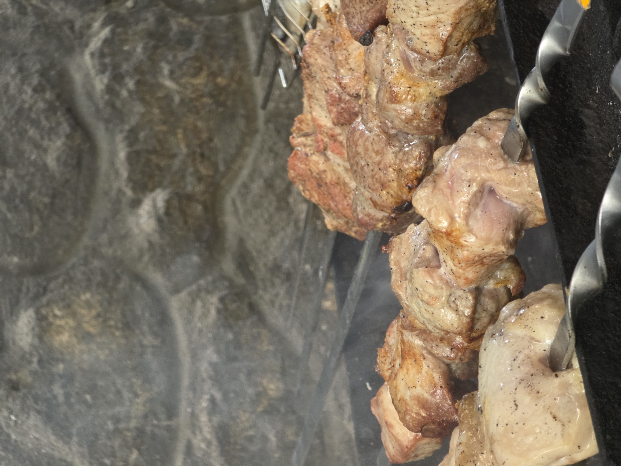 This is a close-up, vertically oriented, eye-level shot of meat grilling on skewers. The subjects are pieces of meat, likely pork, threaded onto metal skewers. The meat is browning and appears to be seasoned with pepper. Smoke is visible rising from the meat, indicating it is actively cooking.
The background on the left is a textured, grey stone surface, possibly the wall of a fireplace or an outdoor grill. The right side of the image shows the dark, textured surface of a grill, with the spiral shape of the skewers clearly visible. The overall scene is one of food preparation, likely a barbecue or cookout.
There are no people visible in the image. The lighting suggests it could be daytime, but the exact time of day is not discernible. The weather is also not indicated. There is no visible text. The location context is Pidhorodne, Ukraine, which suggests this could be a traditional Ukrainian dish being prepared outdoors. The focus is entirely on the food and the grilling process.