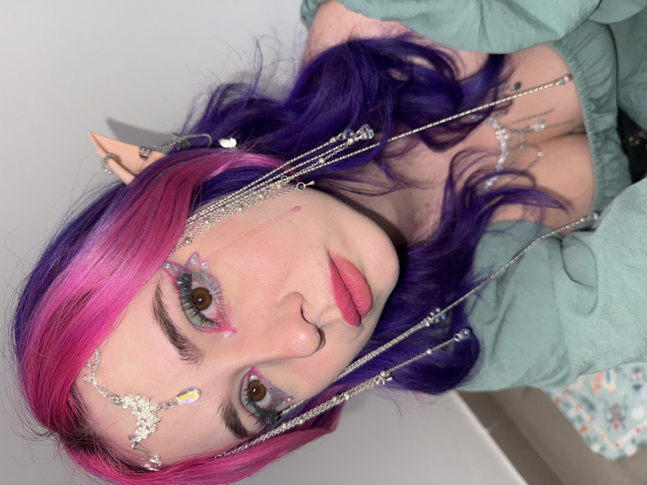 A person with vibrant pink and purple hair, adorned with elaborate silver jewelry around their head and forehead, poses with their head turned. They have pointed elf ears and a light green shirt. Their makeup includes intricate, butterfly-like designs around their eyes and pink lipstick. The focus is on their face, showcasing the fantasy-inspired aesthetic.
