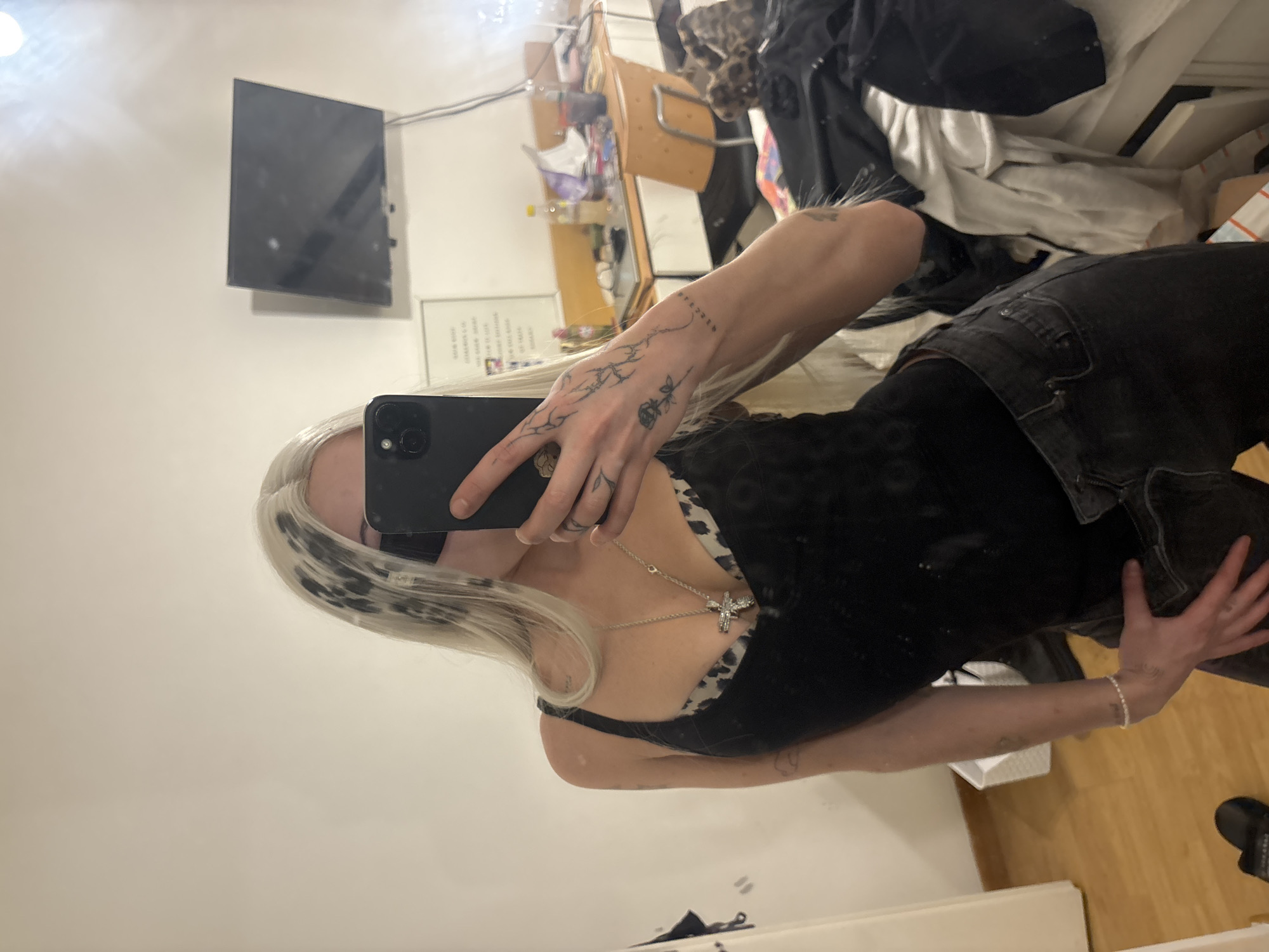 An indoor mirror selfie captures a person taking their own photo. The subject, likely female, has long, light-colored hair with dark roots or streaks, obscuring most of their face with a black smartphone held in their right hand. They are wearing a black top with a leopard print trim, black pants or jeans, and a silver crucifix necklace. Visible tattoos adorn their arms and hands, including intricate line work and a small rose on the right hand, and abstract patterns on the forearms. A pearl bracelet is on their left wrist.
The background, seen in the mirror's reflection, reveals a somewhat cluttered indoor setting, likely a personal room. A wall-mounted flat-screen television is visible on the upper left. To the right of the TV, a wooden cabinet or shelf unit holds various items, possibly toiletries or personal belongings. A pile of clothes or fabric can be seen on a surface behind the person's upper body. The floor appears to be made of light-colored wood. At the bottom right, a black shoe or slipper and a white object (possibly a box or storage bin) are partially visible.
A piece of paper or a sign with text is affixed to the wall behind the subject's arm. Although largely unreadable due to reflection and resolution, some characters are visible, and the word "TODOS" can be faintly discerned. The lighting suggests an indoor setting, but specific time of day or emotions are not discernible. The scene implies a casual moment, characteristic of taking a personal photo in one's living space. While the context provides a Buenos Aires location, no specific visual cues in the image confirm this.