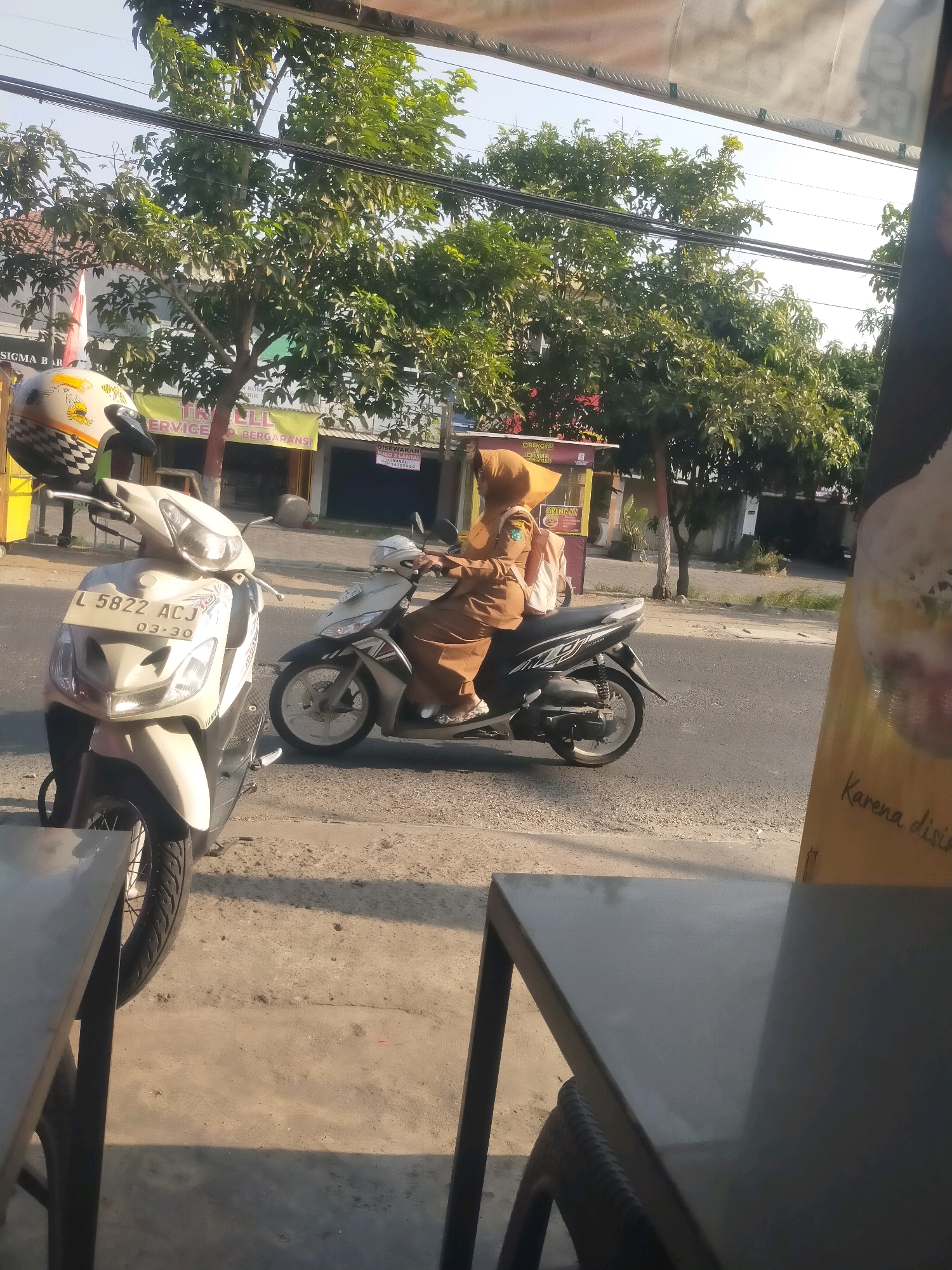 A person wearing a brown hijab and uniform is riding a black motorcycle on a street in Sidodadi, Indonesia. The person is carrying a light-colored backpack. The motorcycle is moving away from the viewer. A white scooter is parked on the side of the road in the foreground. Behind the motorcycles, there are buildings and trees. The sky is visible at the top of the image.