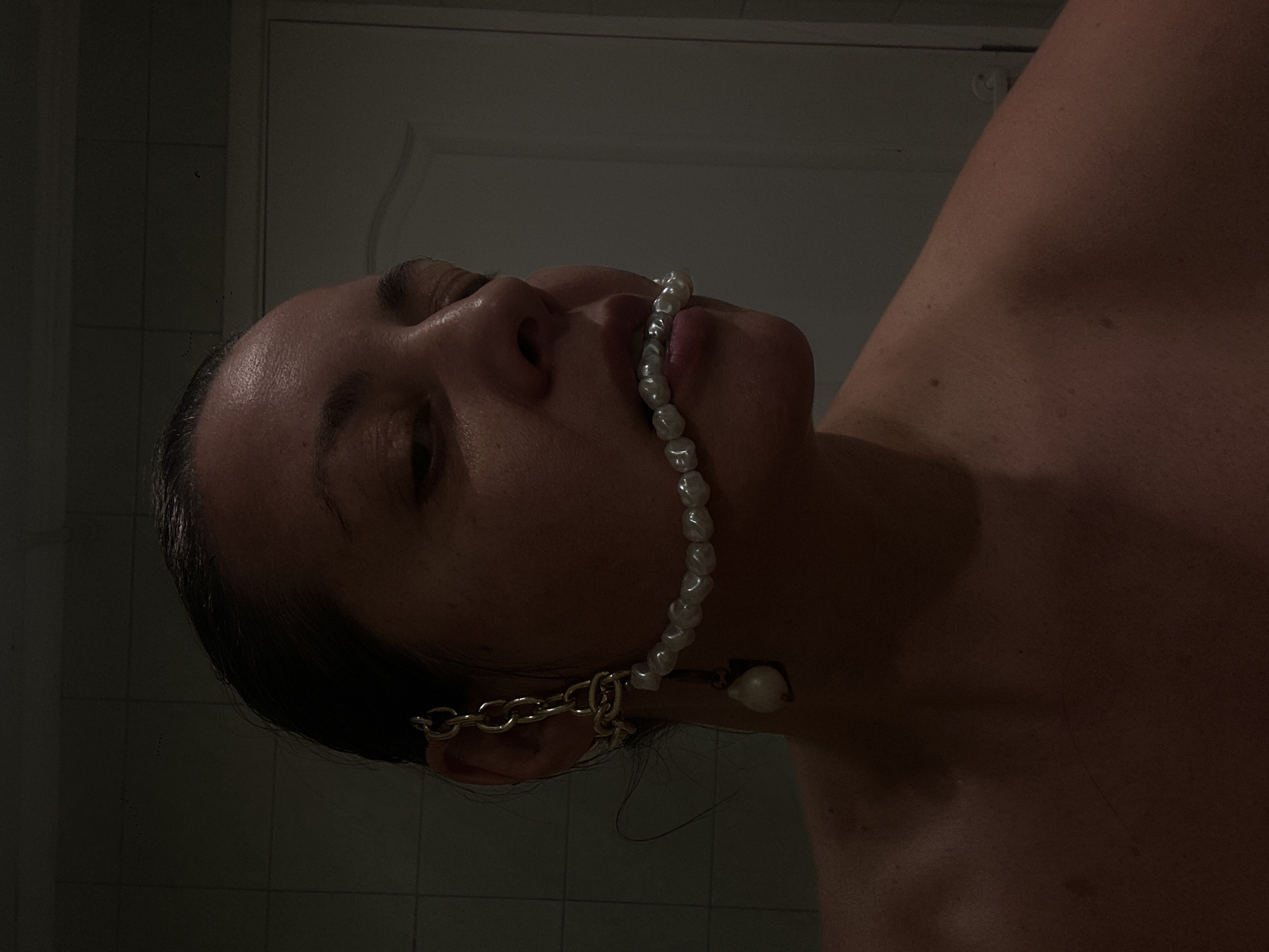 A close-up, low-angle, and slightly rotated image shows a woman with dark hair. She is wearing a string of white pearls in her mouth and a gold chain earring on her left ear. Her eyes are closed, and the background appears to be a bathroom with tiled walls. The location of Riga, Latvia, is not discernible from the visual details provided.
