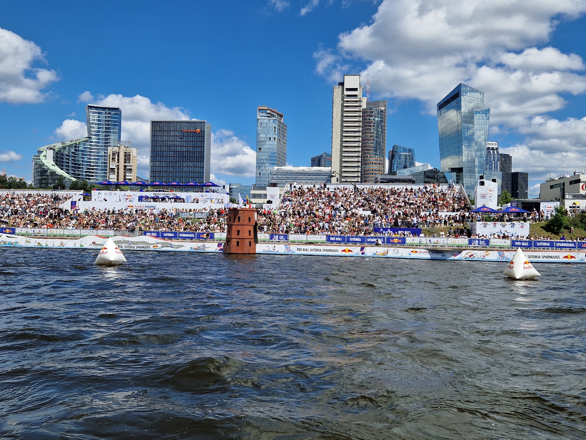The image depicts a daytime scene of a Red Bull event taking place on the Neris River in Vilnius, Lithuania. In the foreground, the dark blue, rippling water of the river occupies the lower half of the frame. A white, triangular buoy with the Red Bull logo is visible on the water's surface to the right.
In the midground, a large crowd of spectators is seated in white bleachers set up along the riverbank. Behind the bleachers, a decorated barrier with the "RED BULL SUTIKIMAS SPARNUIUS" branding runs along the edge of the river. A small, red, brick-like structure, possibly a decorative element or part of the event's setup, stands on the left side of the bleacher area.
The background showcases the modern Vilnius skyline, dominated by several tall buildings. Notable structures include the blue-tinted, wave-like building on the far left, a dark rectangular building with "Swedbank" signage, a tall, slender skyscraper with a glass facade, and other contemporary office buildings. The sky is a vibrant blue, dotted with fluffy white clouds. The overall atmosphere suggests a lively outdoor event in an urban setting.