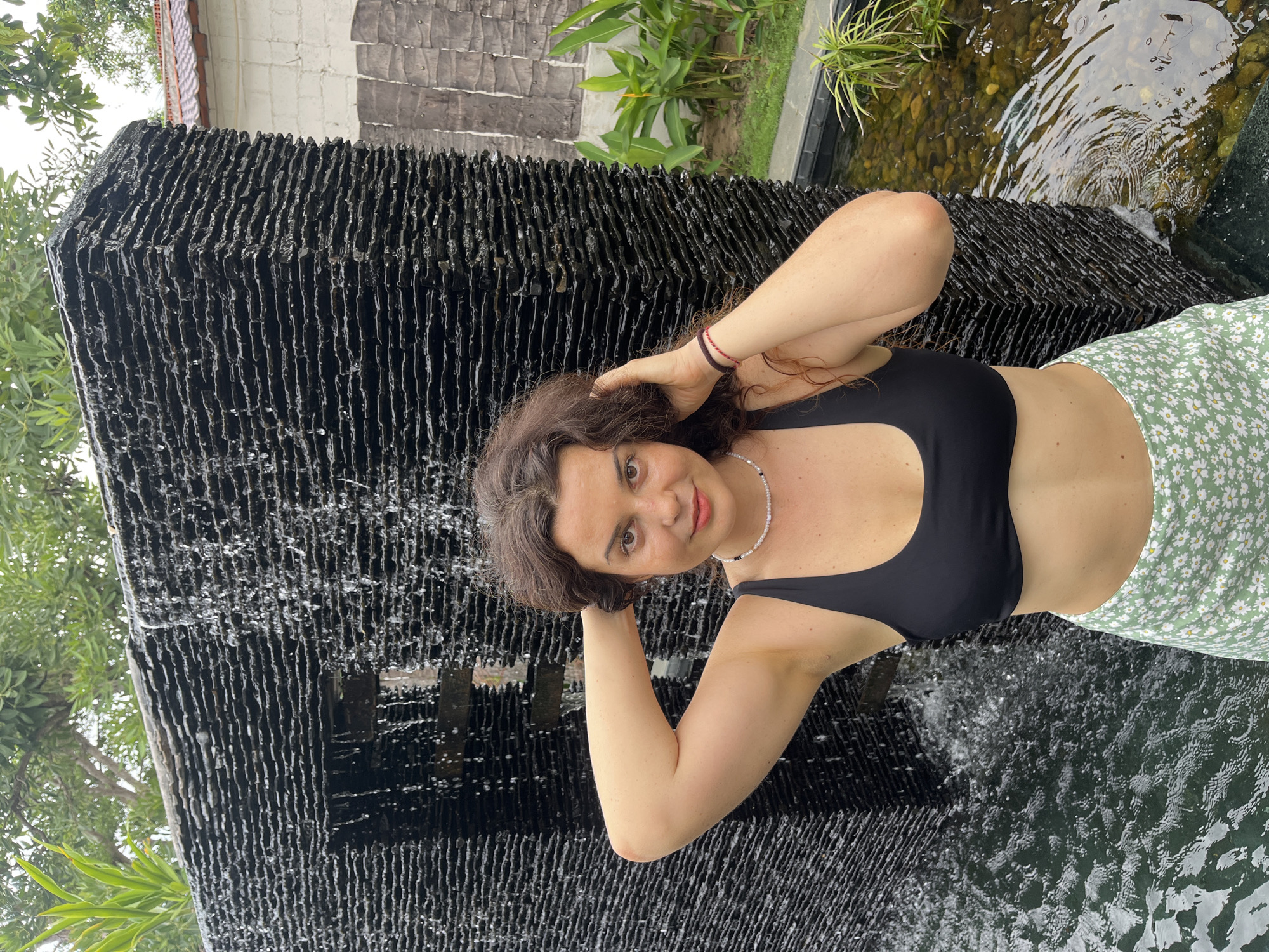 A young woman with curly brown hair and fair skin stands in front of a black, tiered waterfall feature. She is wearing a black bikini top and a green skirt with a white daisy print. Her left arm is raised, with her hand resting on the back of her head, while her right arm is bent at the elbow. Water cascades down the textured black stone in front of her, and a shallow pool with visible stones and aquatic plants surrounds the base of the waterfall. The background shows lush green foliage and a building with a white wall and a red-tiled roof, suggesting an outdoor setting. The overall scene is likely located in Mũi Né, Vietnam, a coastal resort town known for its beaches and natural beauty.