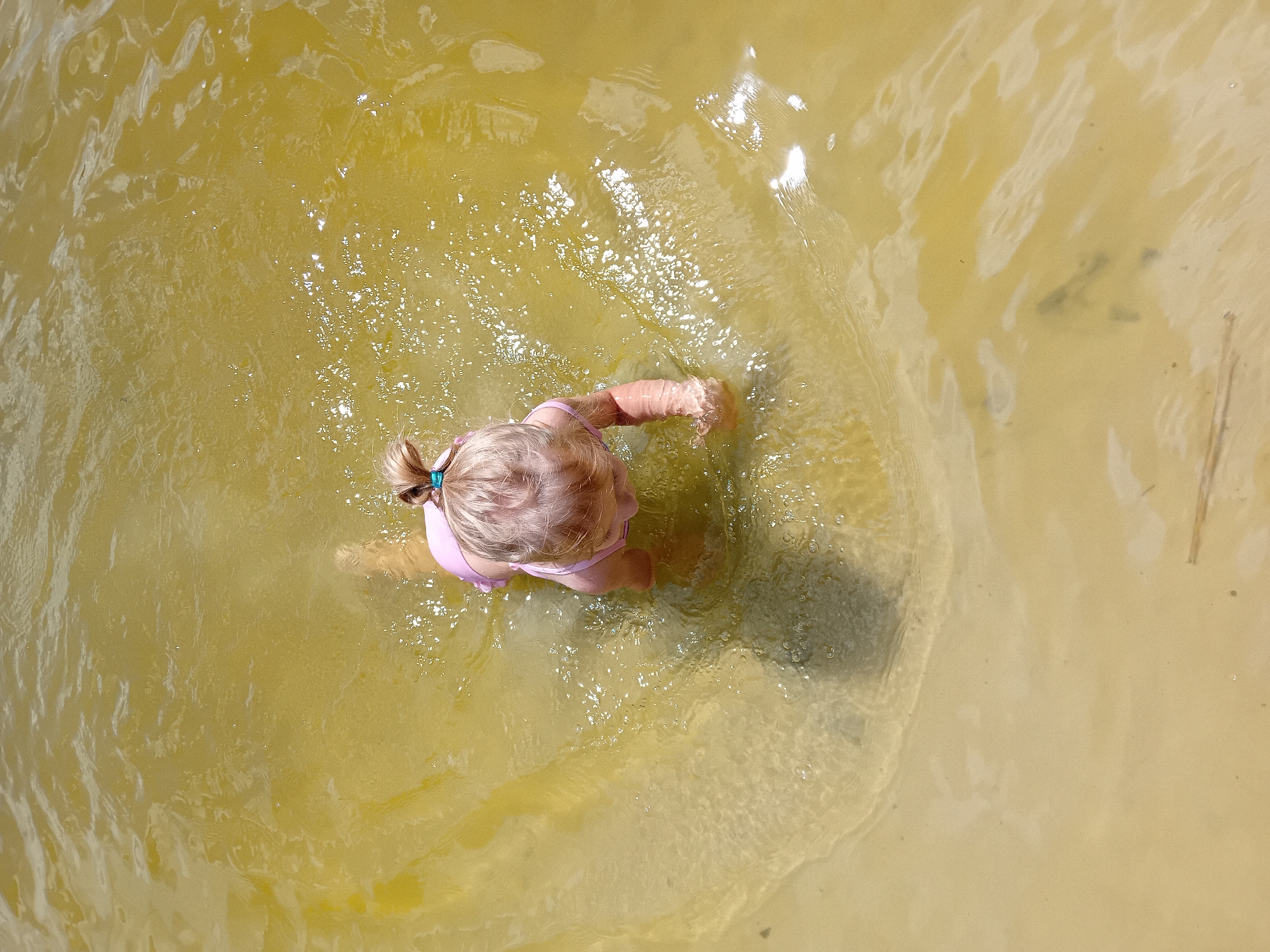 The image is an aerial shot of a young child with blonde hair in a pink swimsuit wading in shallow, murky yellow water. The child is facing away from the camera and appears to be playing in the water, with their hands submerged. The water has a sandy bottom visible in some areas, and there are ripples on the surface indicating movement. Sunlight is reflecting off the water, creating bright spots. The scene suggests a warm, sunny day, possibly in a natural body of water like a lake or river. There are no visible objects or text in the image. The overall mood is one of simple childhood enjoyment in a natural setting. The location context of Chuhuiv, Ukraine, combined with the appearance of the water and the child's activity, suggests a summer day at a local swimming spot.