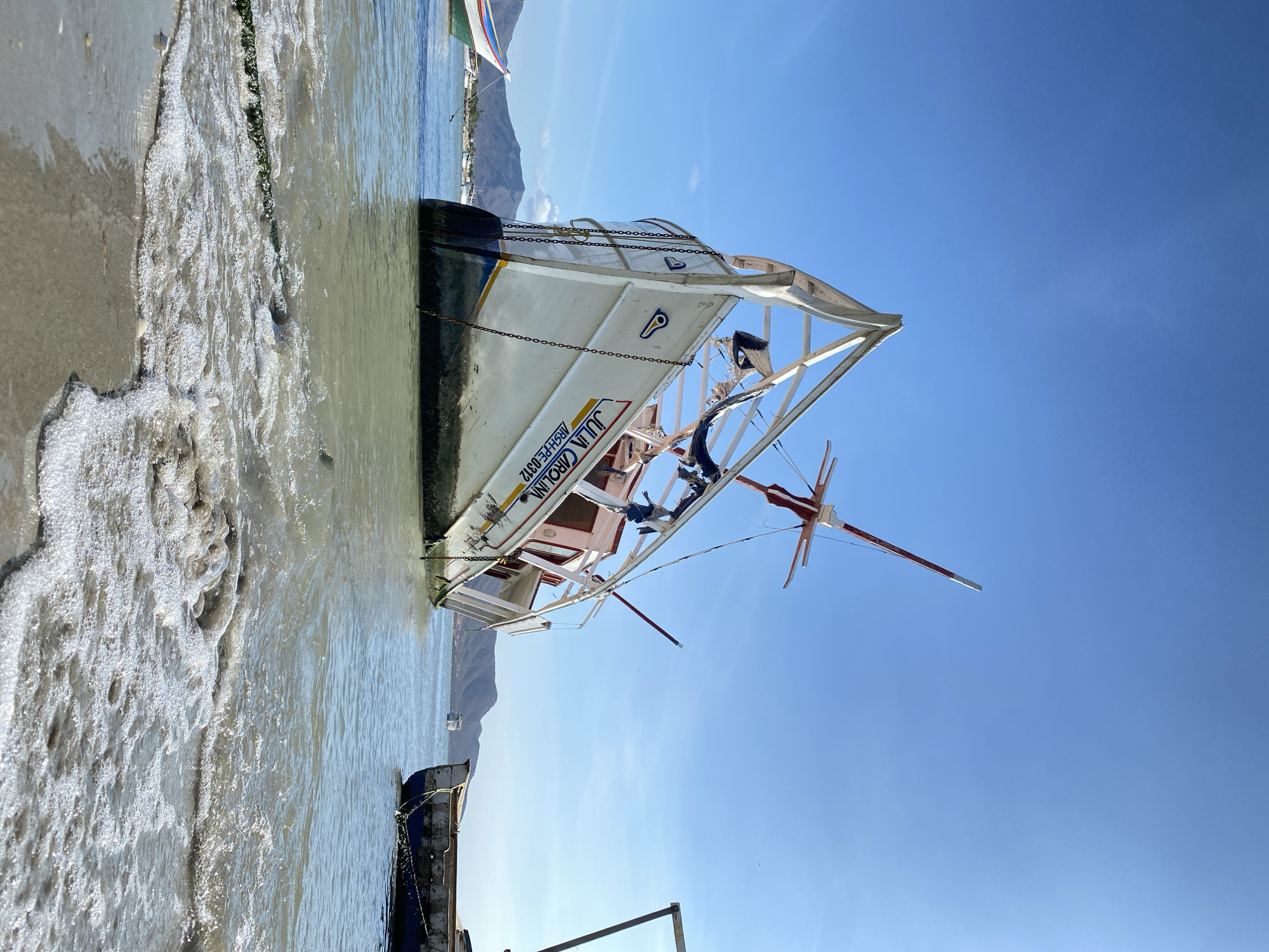 A bright, sunny day on the shores of Juan Griego, Venezuela, is captured in this image, showcasing several boats partially beached in shallow water. The main subject is a white fishing boat, partially resting on a sandy or concrete shoreline, with its bow extending into the murky, light brown water. Foamy waves lap at the shore around the boat.
The boat's hull is dark blue at the waterline, transitioning to white on its upper structure. Chains run along its side. Visible text on the boat clearly reads "JULIA CAROLIN" and below it "ARSHAPE-PE-0312", accompanied by a blue emblem. The boat features an intricate red and white mast or outrigger structure. To the left of this main boat, a smaller, darker vessel, possibly dark blue or black, is also partially submerged.
In the mid-distance, calm, light-colored water extends towards a range of mountains or hills that form the horizon. Another, more colorful boat with green, white, and red stripes is visible in the far background against the mountains. The upper two-thirds of the image are dominated by a vast, clear, vibrant blue sky, indicating excellent weather conditions. No people are visible, lending a peaceful and serene atmosphere to the scene.