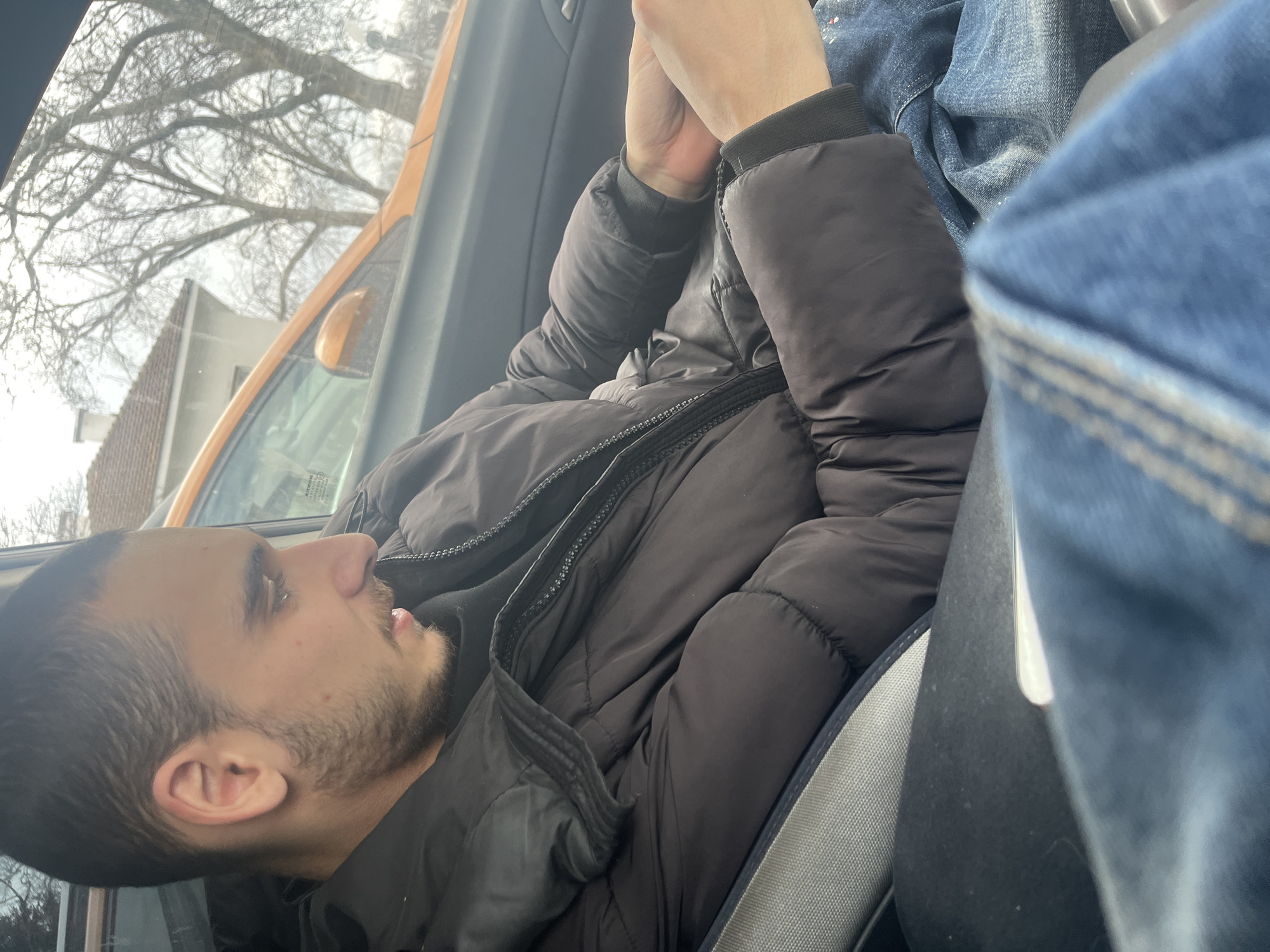 A young man with dark hair and a short beard is seated in a car, wearing a black puffer jacket. He is looking out of the side window. The exterior of the vehicle shows a light orange color. The view through the window includes bare tree branches and a building with a red-tiled roof. The location is presumed to be in or near Varna, Bulgaria. The man's hands are clasped in front of him. Another person wearing denim is partially visible in the seat next to him. The seatbelt is visible.