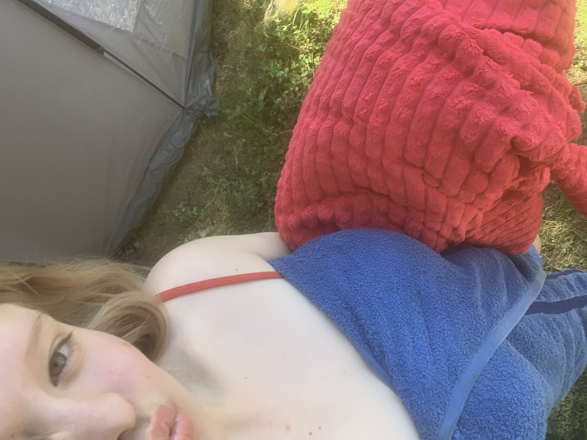 A young individual, likely female, is captured in a close-up, angled shot in an outdoor setting, purportedly in Boitsovo, Russia. Her fair skin, light-colored hair, and a visible hazel eye are prominent on the left side of the frame. Her lips are pursed in a slight pout or kiss, directed towards the camera. She wears a red strap, possibly part of a swimsuit top or camisole, over her right shoulder. A blue terry cloth towel is wrapped around her upper torso and lower body, extending out of the bottom left of the frame. A vibrant red, textured blanket or plush towel is positioned to her right, covering the lower right portion of the image. The ground consists of green grass and visible dirt. In the upper right background, a light grey or tan tent with a darker grey base or ground tarp is partially visible, indicating a camping or outdoor leisure context. The lighting suggests bright daylight. The image itself does not contain specific visual identifiers to confirm the location as Boitsovo, Russia.