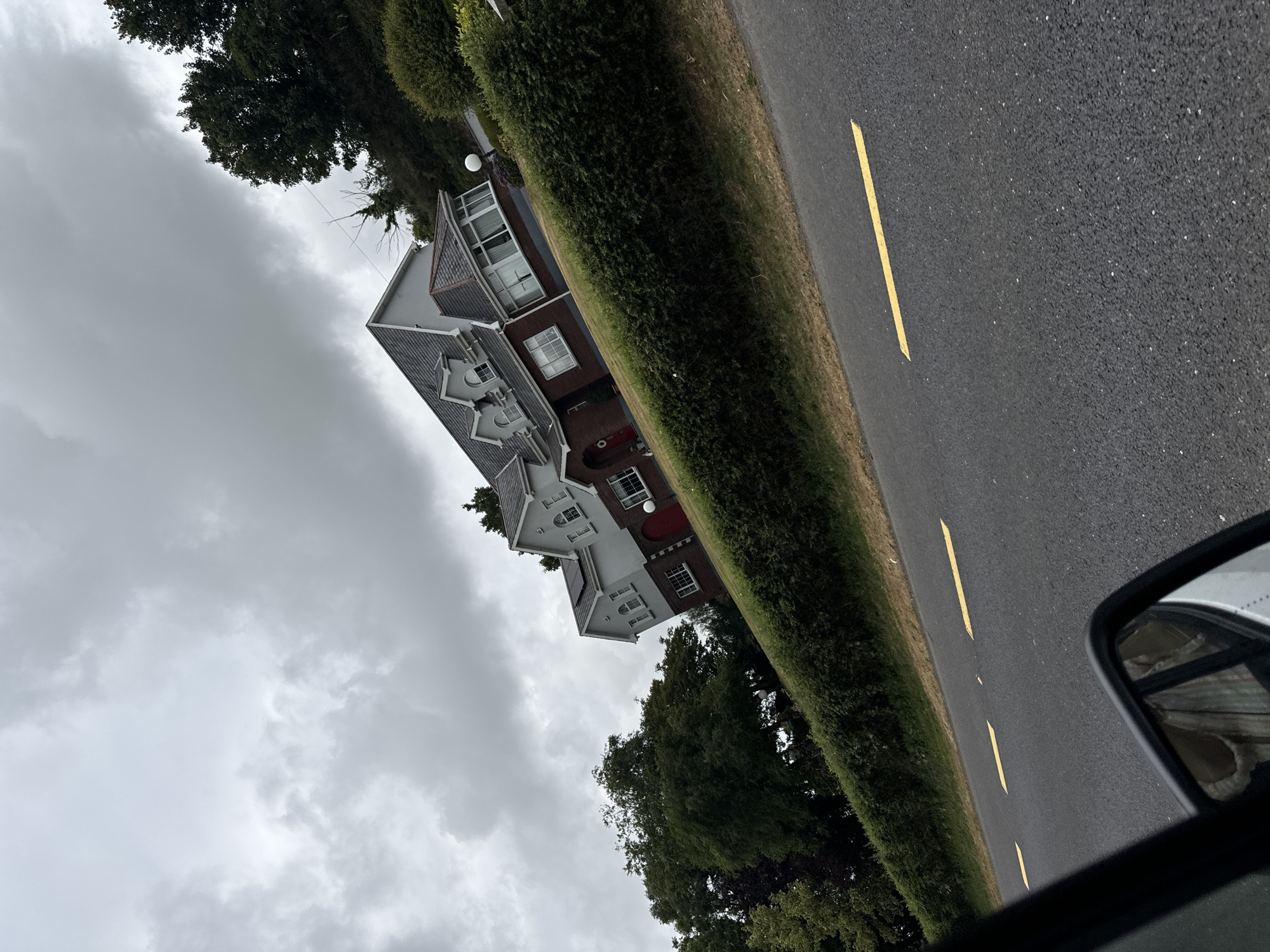 The image depicts a street view of a modern house in the city of Unknown, Ireland. The house is a two-story structure with a gray facade and a dark-tiled roof. It features multiple windows, some with arched tops, and a red front door. The house is surrounded by neatly trimmed green hedges and mature trees. A paved road with yellow lane markings runs in front of the property. The sky is overcast with gray clouds, suggesting a cloudy day. The photograph is taken from the perspective of someone inside a vehicle, as indicated by the side-view mirror visible in the foreground, which reflects a person wearing a patterned garment.