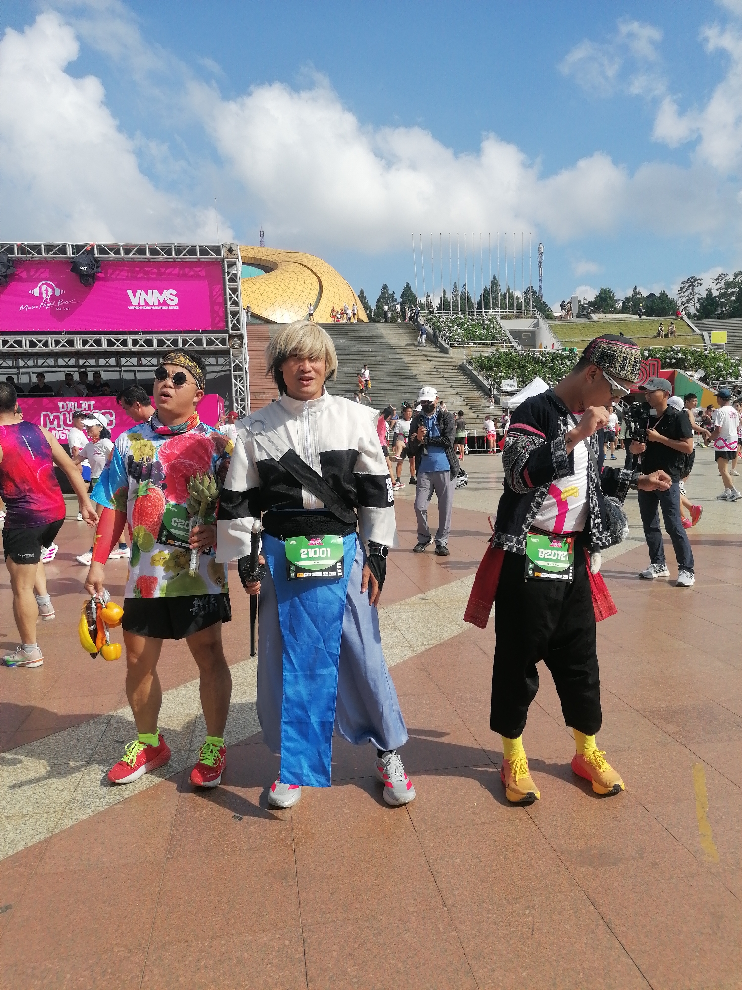 The image depicts an outdoor event at Lam Vien Square in Da Lat, Vietnam, under a clear blue sky with scattered white cumulus clouds. The foreground features three individuals, likely participants in a marathon or race, indicated by the race bibs and banners.
The central-left figure is a light-skinned male wearing dark sunglasses, a patterned bandana, a vibrant short-sleeved shirt with a multi-fruit pattern (including strawberries, watermelon, and lime slices), black athletic shorts, and red running shoes with neon green laces and socks. He holds a cluster of yellow and orange fruits (resembling bananas and persimmons) in his left hand and a single green artichoke in his right. A race bib, partially obscured, is visible on his chest. He looks upwards and to his right.
The central figure is a light-skinned male with shoulder-length, shaggy blonde hair. He wears a costume consisting of a white and black asymmetric long-sleeved jacket over a blue wide-legged lower garment resembling a hakama, worn over dark pants. He holds a sword-like prop in his left hand. A race bib labeled "21001" is attached to his blue garment. He faces the camera with an open-mouthed expression.
The central-right figure is a male wearing a patterned, dark-colored cap, a black long-sleeved jacket with intricate white and red traditional-style patterns, a white t-shirt with a large pink "7" graphic, black pants, and yellow running shoes with matching yellow socks. A race bib labeled "D2012" is affixed to his jacket. He is positioned with his right arm bent, and his left hand slightly raised, looking towards his left.
In the mid-ground, a large pink banner on a metal truss stage structure displays "Music Night Run DA LAT" and "VNMS VIETNAM NIKON MARATHON SERIES." To the right of the stage, the distinctive golden-domed Da Lat Opera House (Artichoke Bud building) with a central circular aperture is visible. Further to the right, a multi-tiered concrete amphitheater-style seating area, adorned with white flowering plants and evergreen trees, ascends. A line of tall, slender white poles stands against the horizon. Numerous other individuals, many dressed in athletic wear, are scattered throughout the background, walking or standing. One individual to the right of the central-right figure holds a professional camera with a large lens. The ground is paved with rectangular tiles in shades of brown and beige.