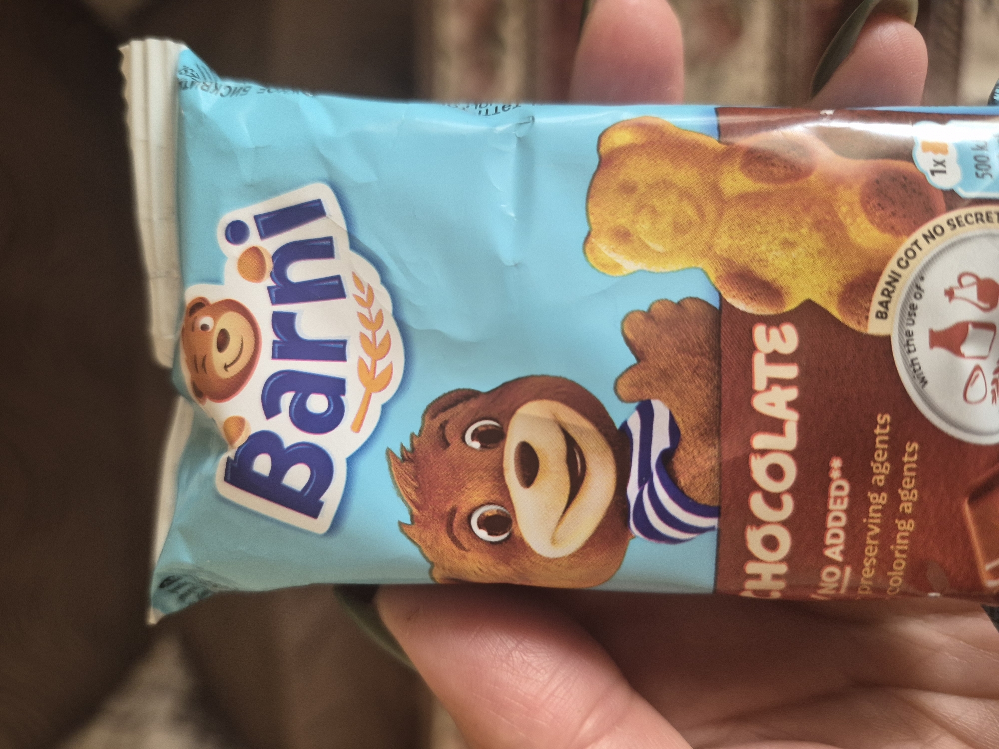 An indoor, eye-level shot captures a hand holding a light blue "Barni" chocolate snack package. The hand, visible in the foreground, has dark green or black nail polish. The package features the prominent "Barni" logo in white and blue, with a small cartoon bear's head. Below this, a larger cartoon bear wearing a blue and white striped shirt is depicted. On the right side of the package, the word "CHOCOLATE" is printed vertically in white text against a brown background.
An illustration of a bear-shaped cake is visible on the upper right side of the packaging. Text elements include "NO ADDED** preserving agents coloring agents" written in white. Partially visible within a circular design is "BARNI GOT NO SECRET with the use of." Further details on the top right show "1x" and "500 k" (likely indicating calories or kilojoules). The background is blurred and dark brown, suggesting an indoor setting. This image is taken in Yerevan, Armenia.