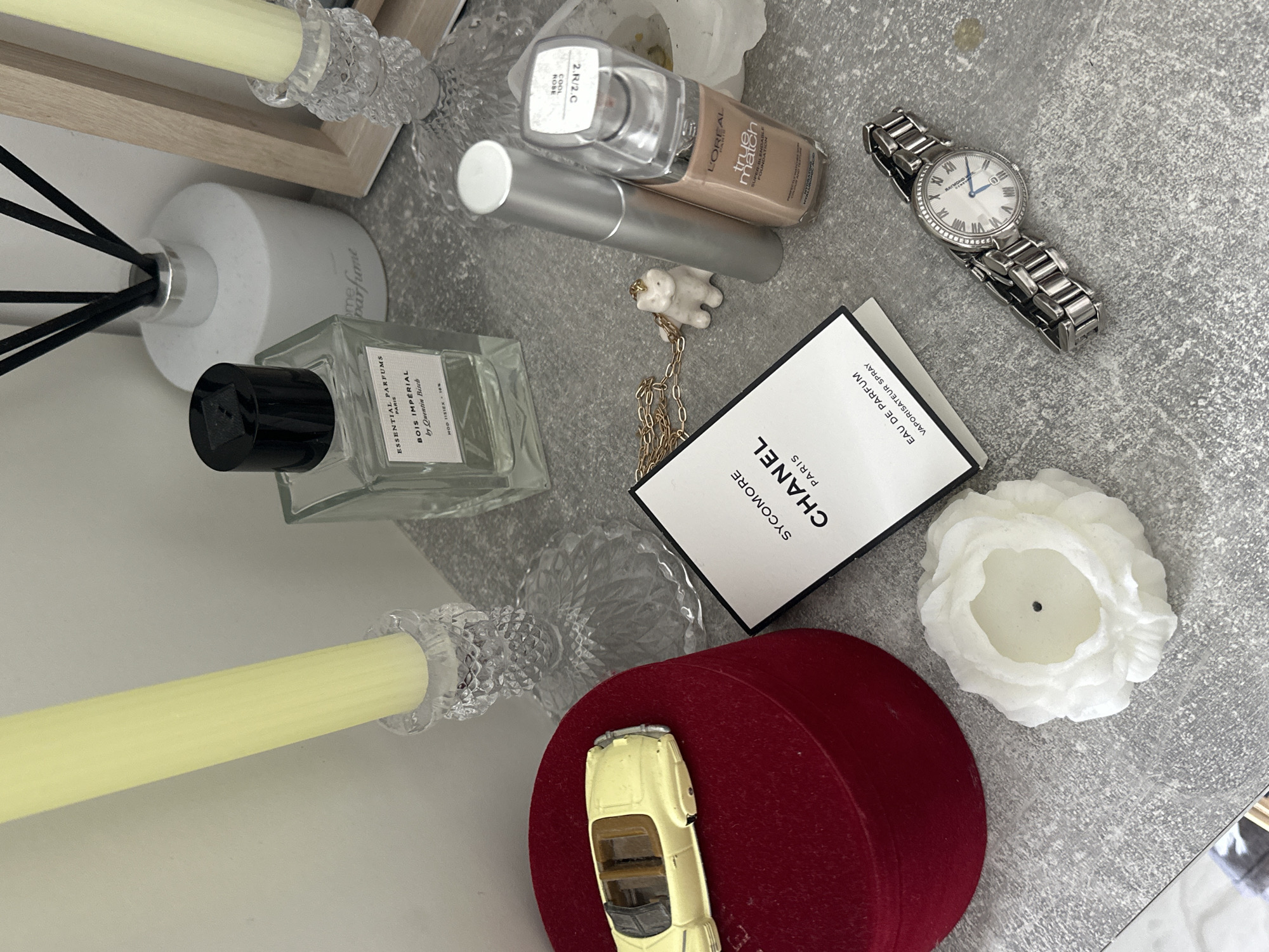 The image is a high-angle shot of a collection of objects arranged on a grey surface, possibly a table or countertop. Dominating the left side are two tall candlesticks holding yellow candles, a bottle of "Bois Imperial" perfume by Essential Parfums, and a white diffuser with black reeds. Moving towards the center, there is a L'Oréal True Match foundation bottle, a silver tube (possibly a concealer), and a necklace with a white bear pendant. A Chanel Sycomore Eau de Parfum box sits beside a white flower-shaped candle. Near the bottom, a red round box holds a yellow toy car. A silver watch with a white face is positioned near the top-right corner. The scene suggests a collection of personal items, potentially related to grooming or aesthetics. There is no indication of the city of Budva, Montenegro, or any other location, people, subjects, or actions.