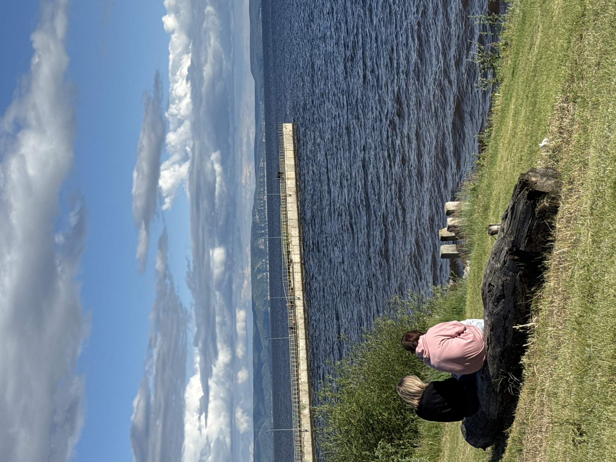 This media file depicts a scene from Primorsky, Russia, likely during daylight hours under a partly cloudy sky. The primary subjects are two individuals, their backs to the camera, seated on a large, dark, weathered log on a grassy embankment. One person is wearing a pink hooded sweatshirt, and the other is wearing a black jacket. They appear to be looking out at a large body of dark blue water, possibly a sea or a wide river.
In the distance, a long, concrete pier extends into the water, with a railing along its side. A few figures can be seen on the pier, suggesting it's a public area. The water is rippling, indicating a gentle breeze. Beyond the water and the pier, a hilly, forested landscape can be observed along the horizon. The sky is a bright blue, interspersed with large, dramatic white and gray clouds, suggesting fair weather. The overall atmosphere seems peaceful and contemplative, with the individuals seemingly enjoying the view and the natural surroundings. There is no visible text in the image.