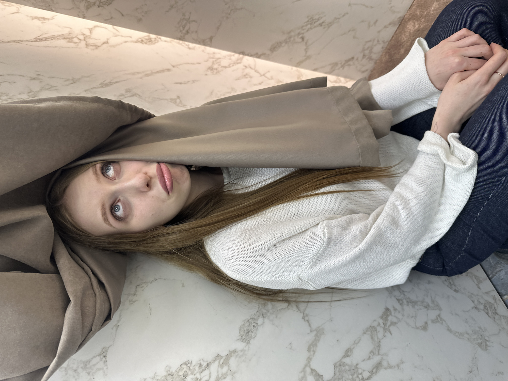 A young woman lies on a marble floor beneath a draped khaki-colored jacket. She wears a white sweater and dark blue jeans. She is indoors; the location is Chaikovsky, Russia.