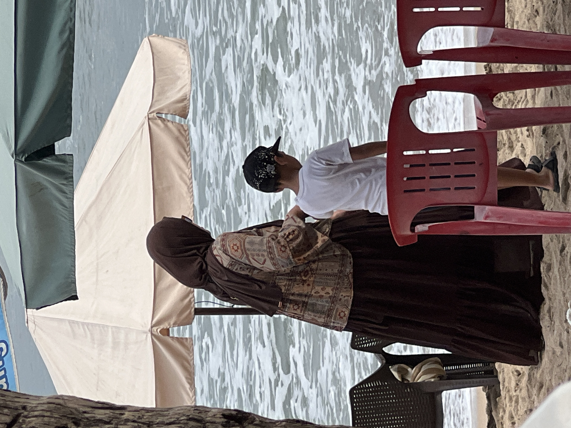 A woman wearing a brown hijab and a patterned tunic over a long brown skirt stands on a sandy beach in Kuta, Indonesia, facing the ocean. Beside her, a young boy in a white t-shirt and a black baseball cap looks out at the water. Red plastic chairs are placed in the foreground, and a dark plastic chair is to the left. Two beach umbrellas, one cream-colored and one dark green, are visible in the background, providing shade from the sun. The ocean waves are white-capped and rolling towards the shore.