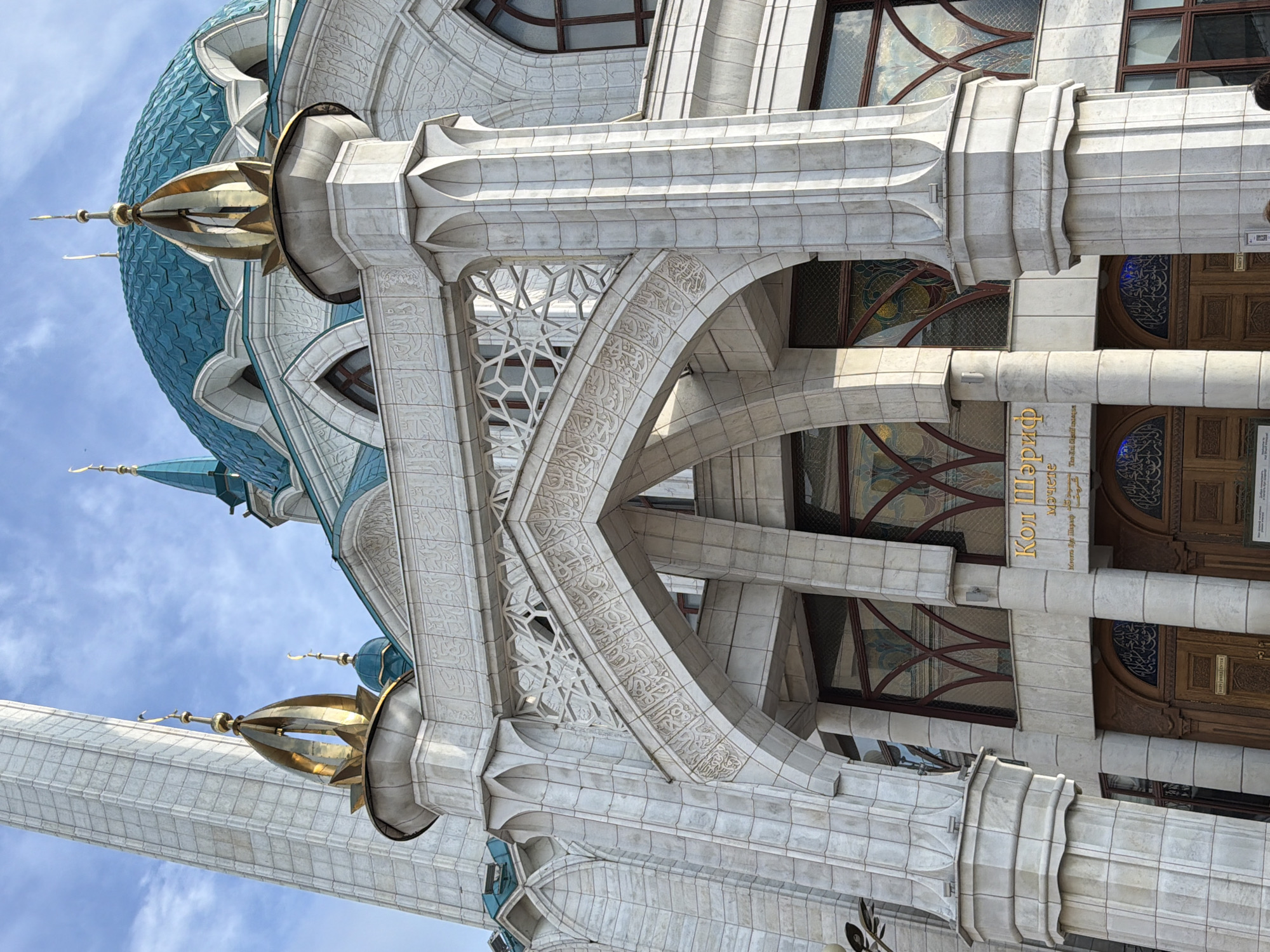 The image shows a close-up, exterior view of a white stone building with intricate architectural details, possibly a mosque given the presence of a minaret and a domed roof structure. The building features elaborate stonework, including decorative carvings, arches, and columns. A tall, slender minaret with a golden finial rises prominently on the left side of the frame. A turquoise-colored dome is visible in the upper left quadrant. The building's facade is adorned with geometric patterns and Arabic script carved into the stone. The windows appear to have stained glass elements.
The lighting suggests it is daytime, with sunlight casting shadows on the facade, indicating a clear or partly cloudy sky. The overall impression is one of grandeur and ornate craftsmanship.
Visible text on the building includes golden lettering in what appears to be Cyrillic and possibly Arabic script. One sign reads "Кол Шәриф", which translates to "Kul Sharif" in Tatar and Russian, a strong indicator that this is the Kul Sharif Mosque in Kazan, Russia, a famous landmark. The text suggests it is a public building, likely a religious or historical site. There are no people clearly visible in this frame. The scene is static, focusing solely on the architectural elements of the building against a sky background.