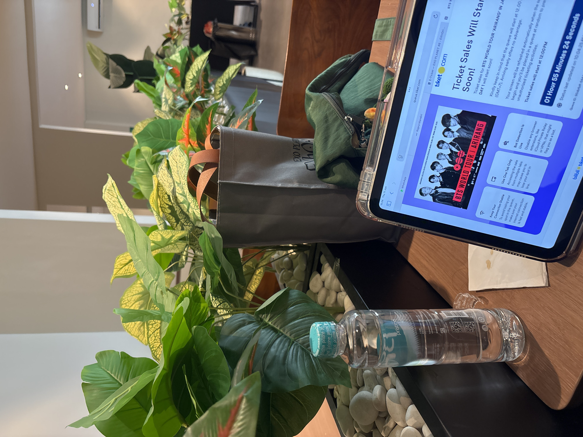 The image depicts a close-up view of a wooden table with various objects. In the foreground, a bottle of "Pristine" water sits next to a crumpled white paper napkin. To the right, a tablet device displays a webpage with a BTS concert advertisement and information about ticket sales. Behind the water bottle, a dark planter box filled with decorative white stones is visible, with artificial green plants emerging from it. A gray fabric tote bag with brown straps stands behind the planter. Further back, more plants and a green pouch with a zipper are visible, along with part of a wooden chair. The overall setting appears to be indoors, possibly a cafe or a home, with a blurred background suggesting a neutral-colored wall and possibly a ceiling light. The webpage on the tablet indicates the ticket sales are for "BTS WORLD TOUR 'ARIRANG'" in Jakarta, Indonesia, specifically at the Jakarta Gelora Bung Karno Main Stadium on December 26, 2026, at 12:00 PM GMT+7. The tablet's system clock shows "10:04 Thu 11 Jun." While the image captures elements related to a concert in Jakarta, the specific location within West Jakarta is not discernible from the visual information provided.