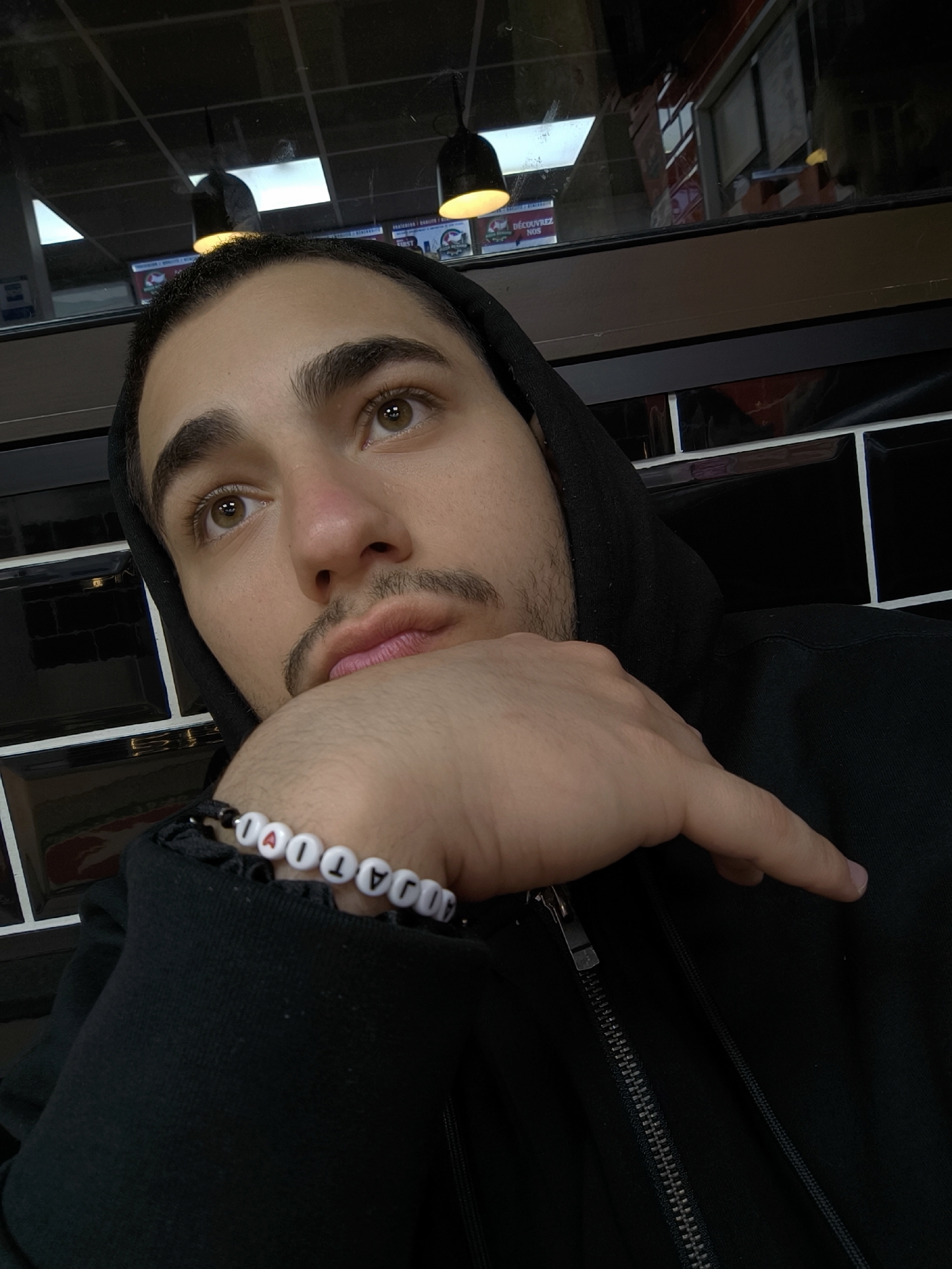 A young man is depicted in an eye-level, close-up shot, seemingly indoors. He is wearing a black hooded sweatshirt. A bracelet is visible on his wrist, containing letter beads spelling out "AJATIOLA". The man is resting his head on his hand, gazing upward and leftward with a contemplative expression. Behind him are dark, tiled walls, likely an interior. Through a reflective window above, light fixtures and exterior signage in French can be observed, pointing to Strasbourg, France. There is no discernible action or interaction taking place. The environment consists of the interior space with reflections of the outside.
