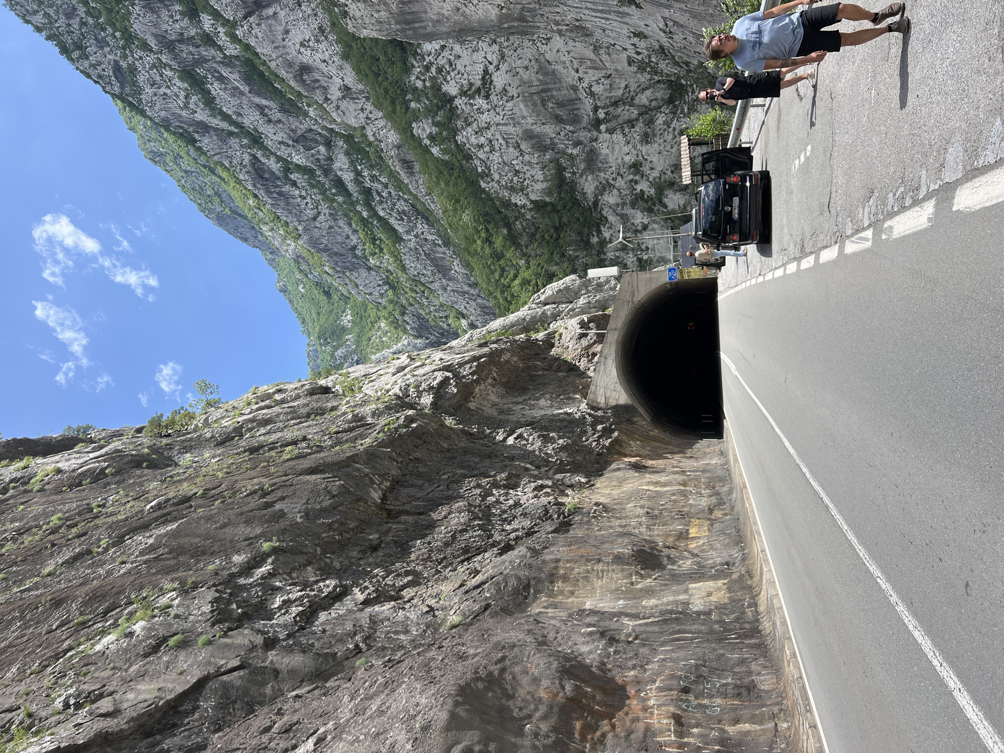 The image shows a road in Vlahovići, Montenegro, leading into a tunnel carved into a rocky mountain. A black car is parked on the side of the road near the tunnel entrance. Two people stand on the road near the car: a man with sunglasses and shorts, and another person taking a picture. The mountain rises steeply on both sides of the road, and the sky is visible above.
