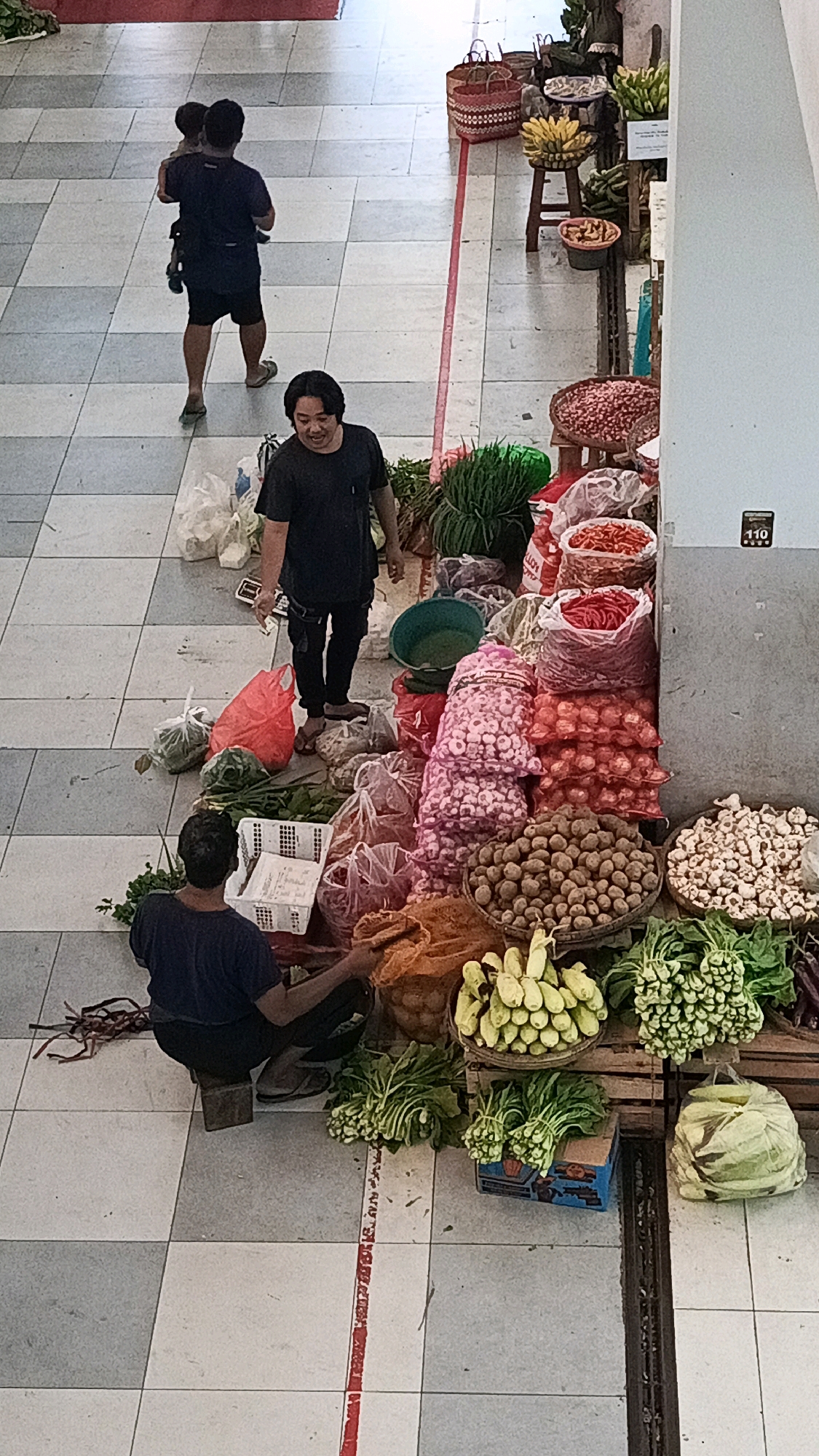 The image depicts an outdoor market scene in Ponorogo, Indonesia, with a focus on fresh produce. In the foreground, two men are visible. One man is squatting and sorting vegetables, while another man, wearing a dark t-shirt and dark pants, stands near a display of produce, holding a cigarette and a piece of paper. In the background, a person is carrying a child. Various types of vegetables, including leafy greens, onions, potatoes, and chili peppers, are displayed in piles and bags. Woven baskets and crates are used for storage and display. The market floor is tiled with a grey and white checkered pattern. A red line is painted on the floor, possibly indicating a boundary or pathway. A sign with the number "110" is affixed to a wall on the right side of the frame. The overall scene suggests a vibrant and active marketplace.