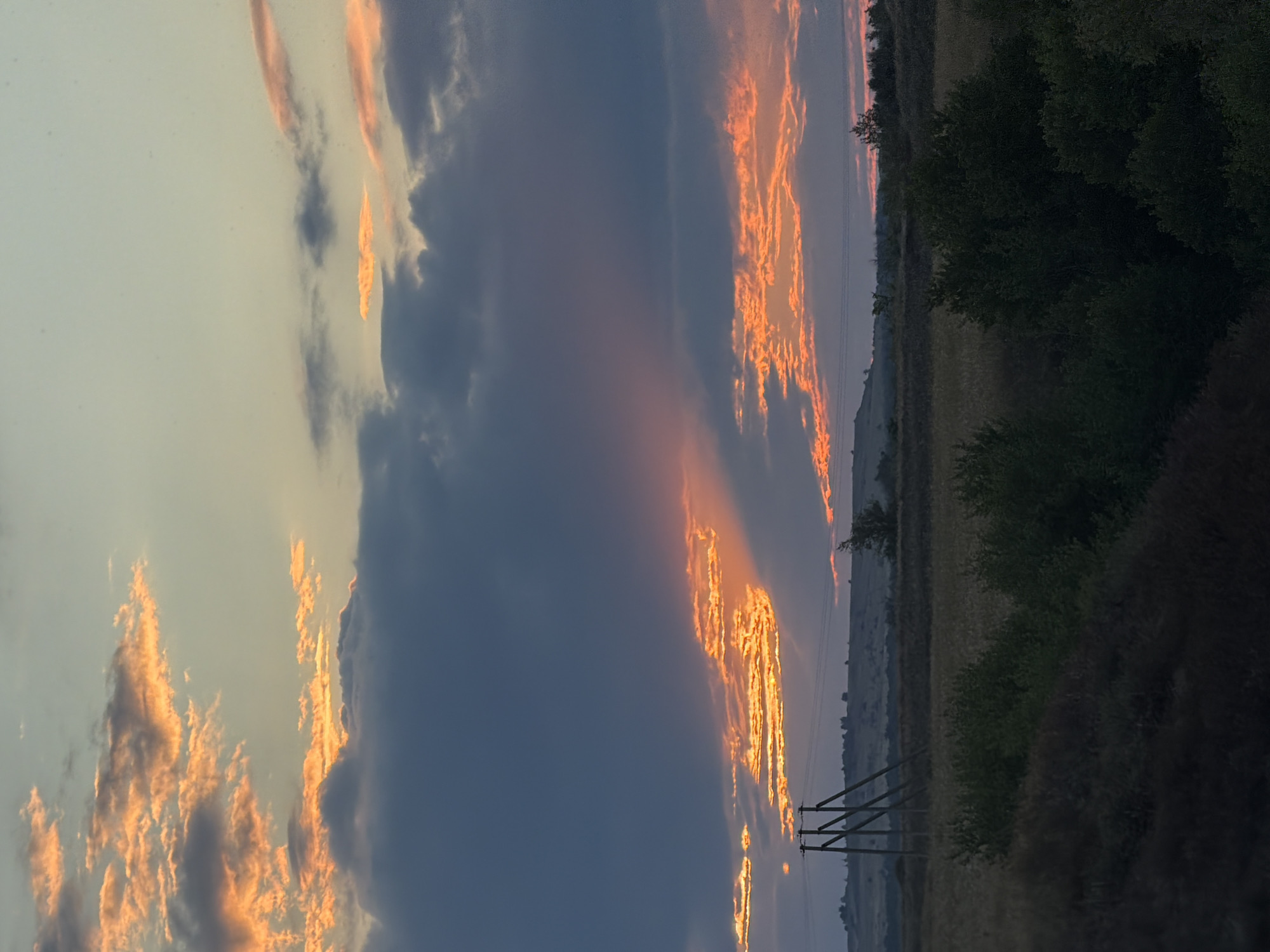 A vertical, eye-level medium shot captures a vibrant sunset over a rural landscape. The sky is dominated by dramatic clouds, with patches of orange and pink light breaking through darker, gray formations. The light creates a sense of depth, with sunbeams fanning out from behind the clouds. In the middle ground, a power line tower with wooden crossbeams stands to the left, silhouetted against the horizon. Beyond the tower, a flat expanse of dry grass or crops stretches to a gently rolling, distant hill. The foreground is a dark, silhouetted band of vegetation, with some lighter green bushes and trees interspersed throughout the scene. The overall impression is one of a natural, tranquil evening in a rural setting near Nizhnyaya Lipovka, Russia.