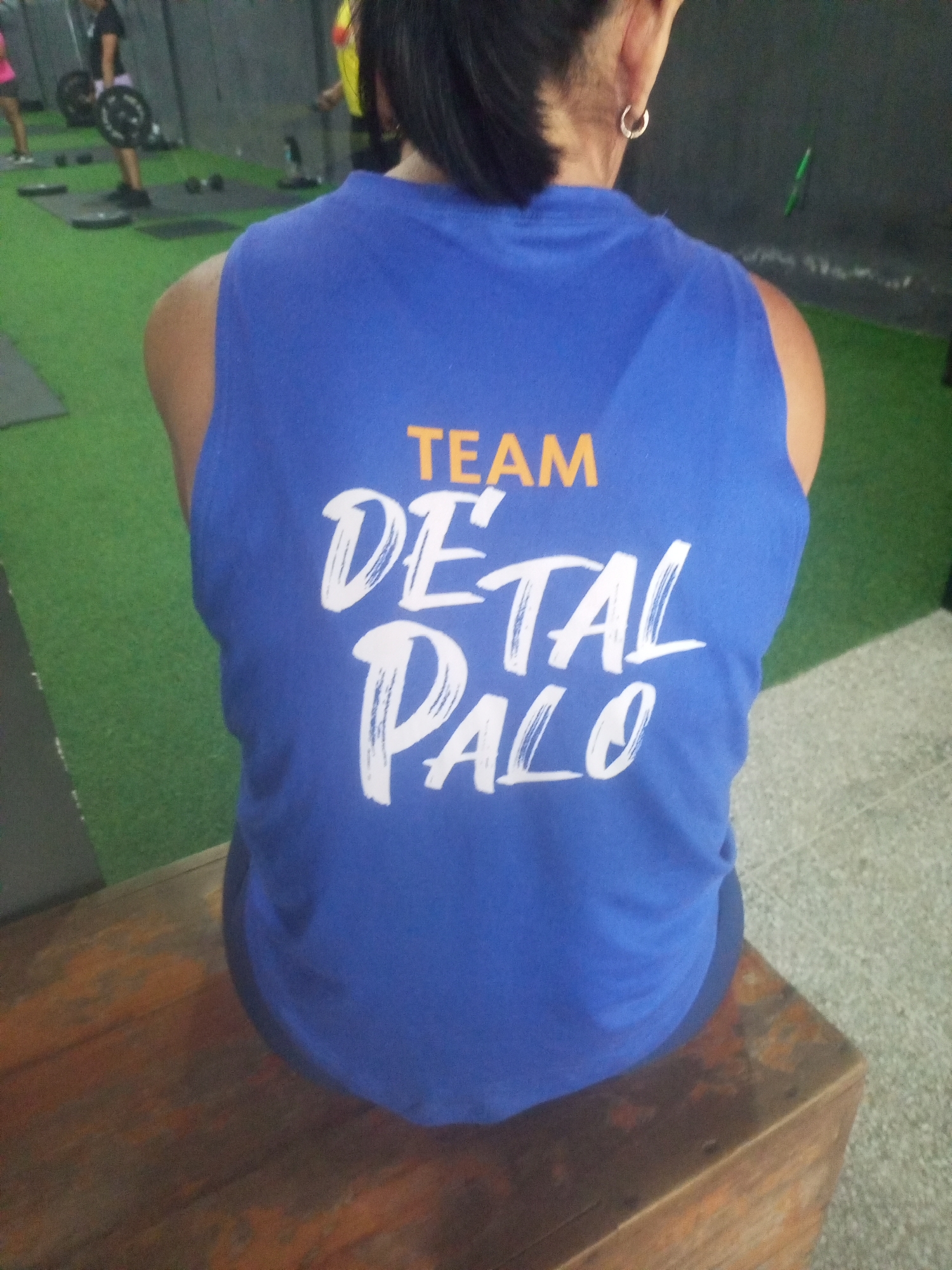 A woman wearing a blue tank top with the text "TEAM DETAL PALO" printed on it in orange and white is seated on a wooden bench in a gym. She is viewed from behind, and her back is towards the camera. The gym floor appears to be a green turf, and there are various pieces of exercise equipment visible in the background, including weight racks and dumbbells. The lighting in the gym is dim. The overall scene suggests a casual workout setting.