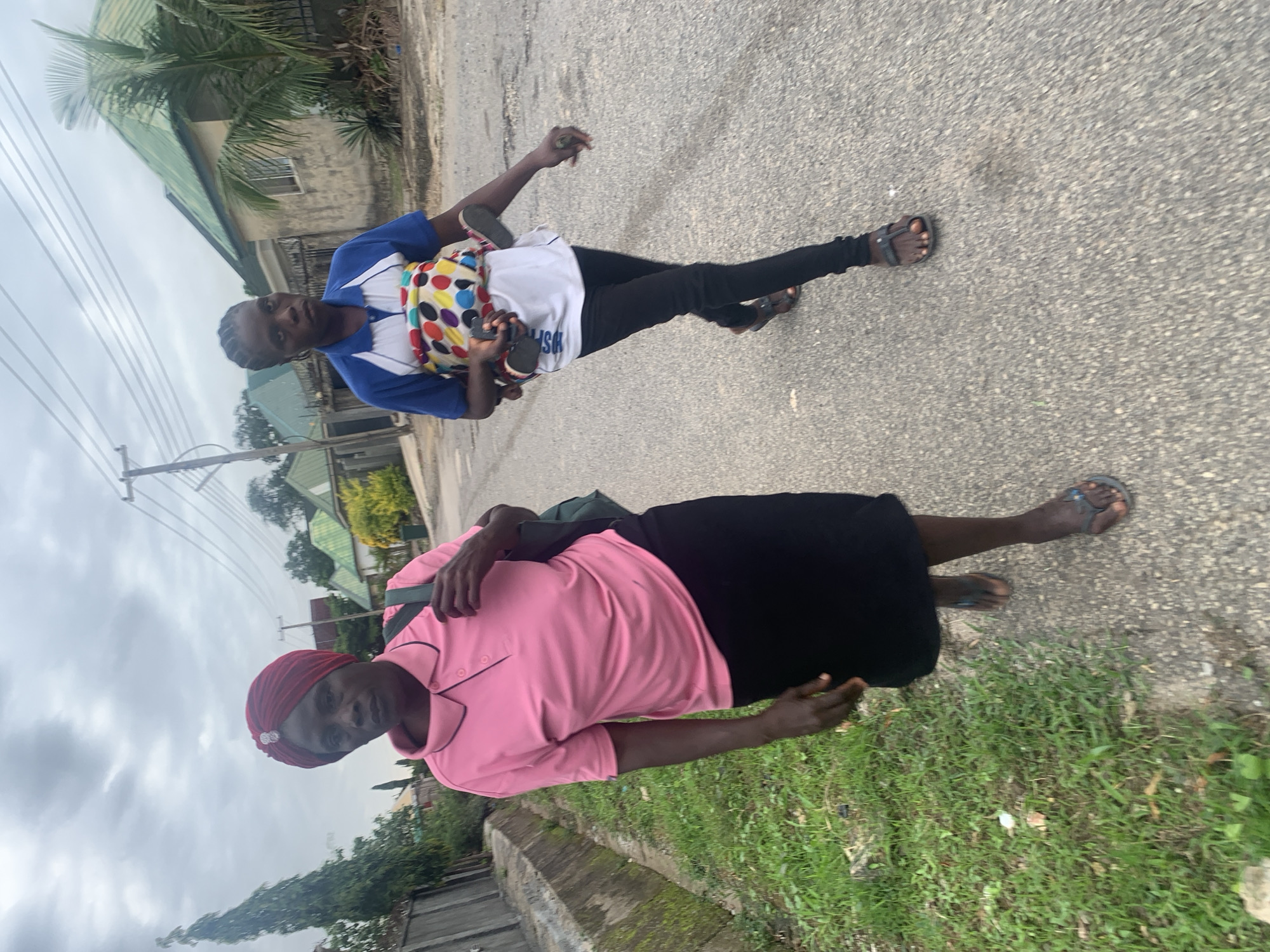 Two women are walking on a paved road in a residential area of Abuja, Nigeria. The woman in the foreground is wearing a pink polo shirt, a black skirt, and flip-flops. She has a maroon headscarf on and a dark bag slung over her shoulder. The woman in the background is wearing a blue and white polo shirt with a colorful baby carrier wrapped around her. She is wearing black pants and flip-flops. The houses in the background appear to be single-story structures with green roofs. The sky is overcast and the lighting is dim. There is grass growing along the side of the road and a concrete drainage ditch. Power lines are visible overhead.