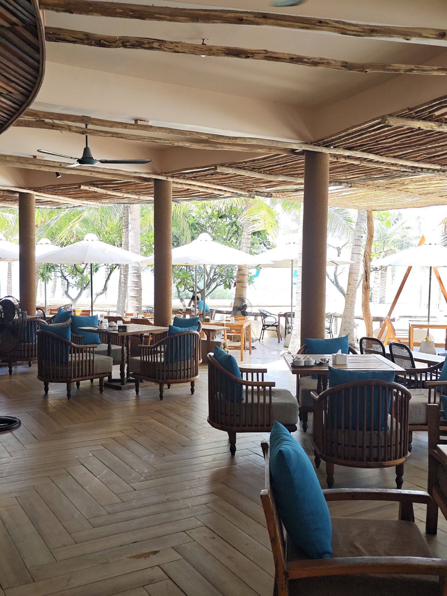 A beachfront restaurant in Phan Rang – Tháp Chàm, Vietnam, features an open-air dining area with a thatched roof supported by thick, rope-wrapped columns. The floor is made of light-colored wooden planks laid in a herringbone pattern. Rows of seating areas with wooden chairs and tables, adorned with teal cushions, are arranged throughout the space. White umbrellas are positioned outside, suggesting a sunny day. Lush green palm trees are visible beyond the seating area. A ceiling fan is mounted above the dining area.