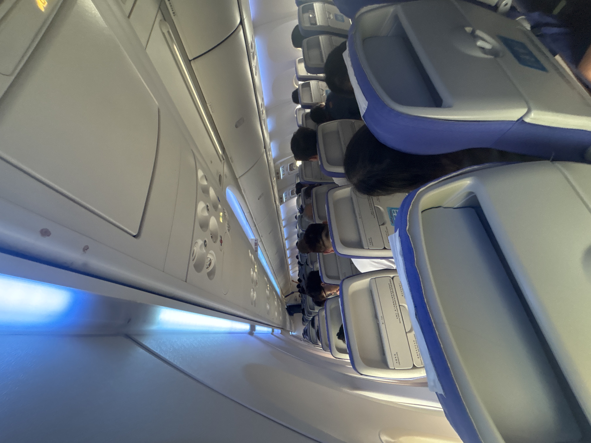 The image shows the interior of an airplane cabin, likely a commercial airliner. The perspective is from a passenger's seat, looking forward down the aisle. The seats are upholstered in a light gray fabric with blue accents. Overhead bins are visible, as are the overhead reading lights which are illuminated with a cool blue glow. Several passengers are visible in their seats, mostly with their backs to the camera, suggesting the plane is either boarding or in flight. The image is taken in Baiyun District, China, which is home to Guangzhou Baiyun International Airport, a major hub for air travel.