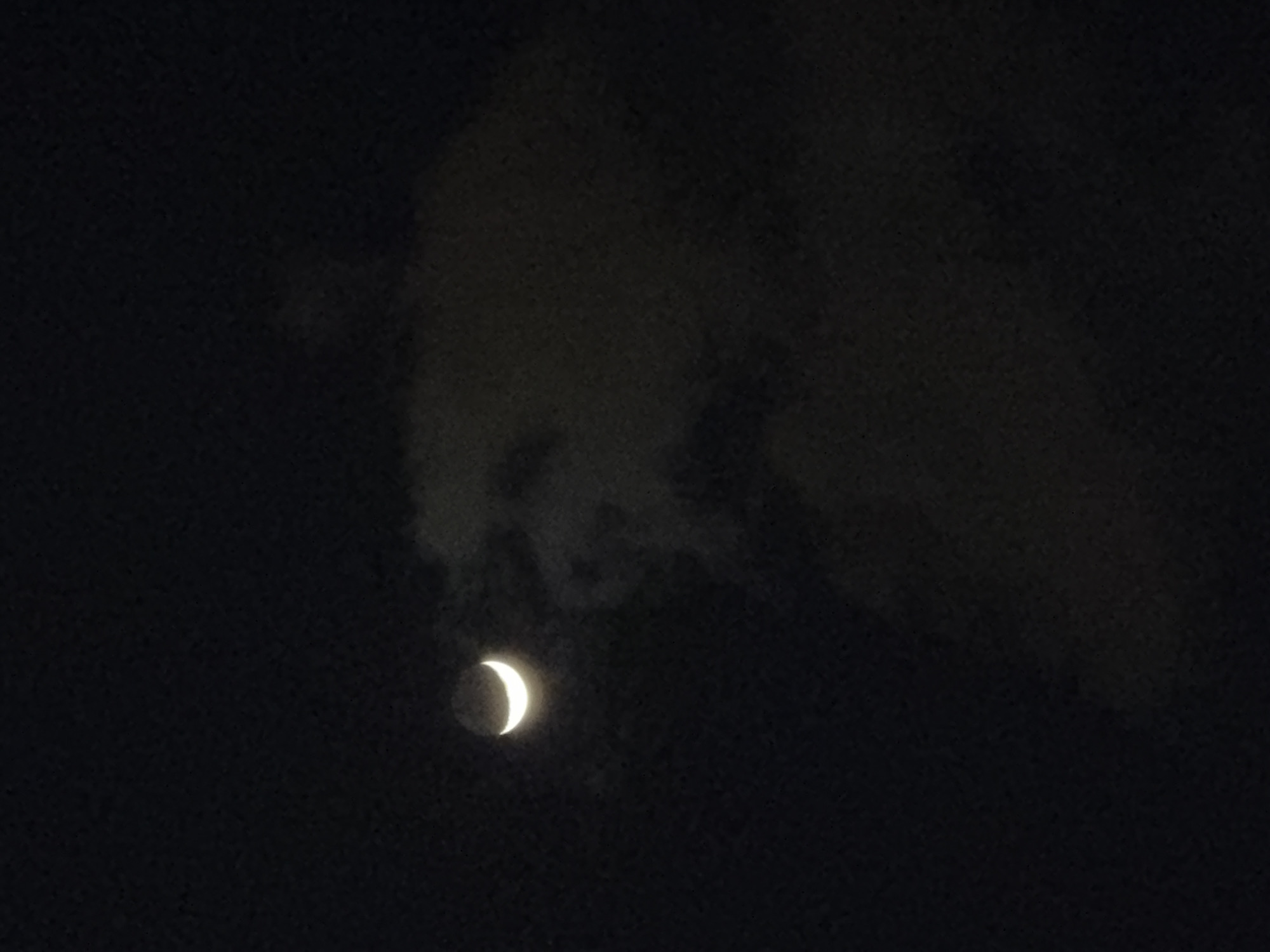 The media file presents a clear image of a crescent moon in a dark night sky, likely captured over Kiryat Bialik, Israel. The bright, illuminated portion of the moon is sharply defined, exhibiting a distinct crescent shape with a soft white-yellow glow. Dark, amorphous clouds are scattered across the upper and central parts of the frame, partially obscuring the sky around and above the moon, lending a textured quality to the otherwise deep black background. The clouds appear to be thin and wispy in places, revealing subtle variations in their density against the darkness. The image is focused on this celestial phenomenon, with no other discernible objects or figures present. The time of day is clearly night. No specific weather details beyond the presence of clouds can be inferred, and no text is visible. The scene evokes a sense of calm and natural beauty.