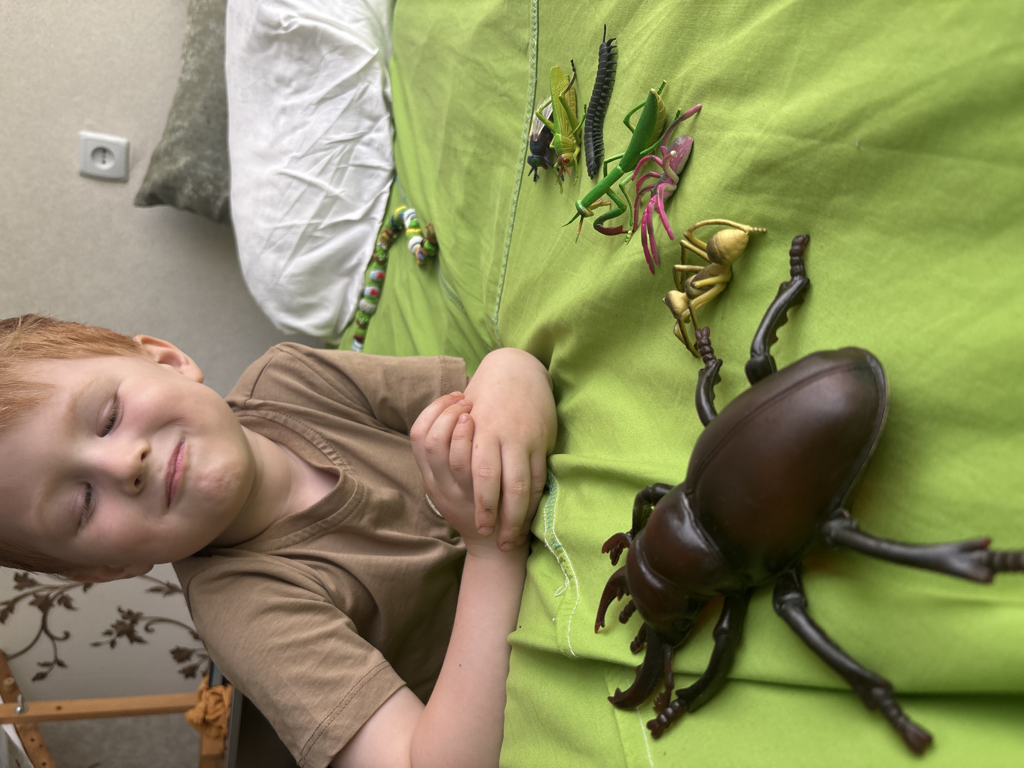 A young, light-skinned, red-haired child lies on a bed covered with bright green bedding. The child has their eyes closed and a slight smile. Lying on the bed with the child are several plastic insects, including a large, brown rhinoceros beetle and several mantises of different colors. A stuffed toy caterpillar is also visible on the bed. An electrical outlet is visible on the wall. The location is Russia, in the city of Chaikovsky.