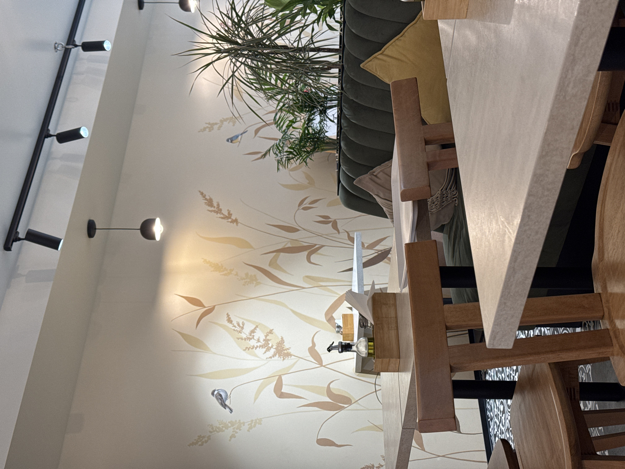 An interior shot of a restaurant or cafe in Budva, Montenegro. A green velvet booth with yellow pillows sits against a wall decorated with a wheat pattern and small birds. A table is close to the camera. A wooden stand with oil and vinegar bottles is visible on the wall. Track lighting runs along the ceiling. The image is rotated counter-clockwise.