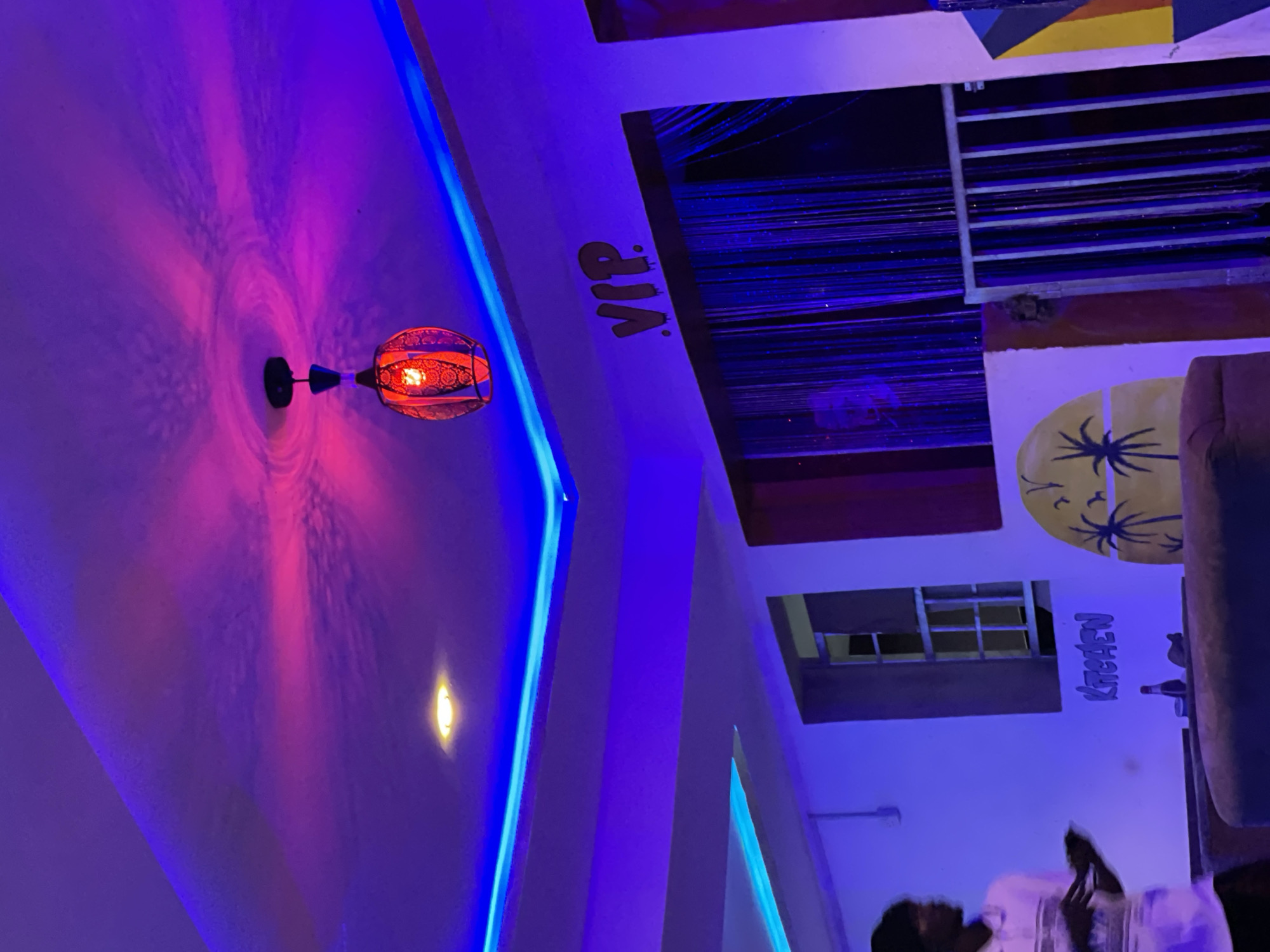 This is a medium shot, eye-level, indoor photograph taken in the city of Durumi, Nigeria. The scene appears to be a dimly lit establishment with purple and blue ambient lighting, likely a lounge or bar. Dominating the upper portion of the image is a ceiling illuminated by blue LED strip lighting along its edges, casting a violet glow on the textured ceiling itself. A hanging lamp with a red, intricate glass shade and a visible light bulb is suspended from the center of the ceiling, projecting a pinkish-red light pattern onto the ceiling. A small, recessed yellow light is also visible on the ceiling.
On the wall, the word "VIP" is spray-painted in a dripping font above a curtain of shimmering, dark vertical strands, possibly beaded or sequined. To the left of the "VIP" sign and curtain, a rectangular window with a metal grate is visible. Below the window, the word "KITCHEN" is spray-painted in a similar style to "VIP," with a bottle and another object resting on a surface below it. To the right of the "KITCHEN" sign, a circular mural featuring a palm tree silhouette against a yellow background is present.
In the foreground, a person wearing a light-colored collared shirt with a patterned design is standing on the left side of the frame, facing towards the right. They appear to be holding a dark object in their right hand and looking down. The lower part of the image shows the tops of dark upholstered furniture, possibly couches or seating. A metal railing or gate is visible to the right of the beaded curtain. The overall ambiance is moody and suggestive of a nightlife venue.