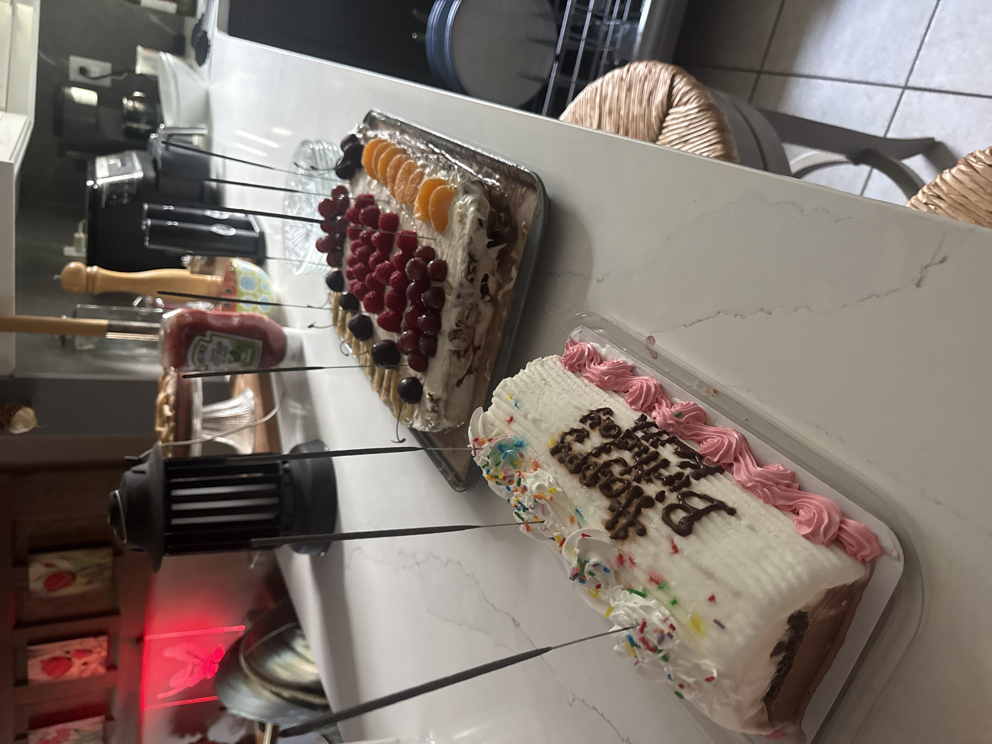 Two birthday cakes with sparklers are displayed on a white countertop. The cake in the foreground is a rolled cake decorated with white frosting, colorful sprinkles, pink piped frosting along the edges, and chocolate lettering that reads "Happy Birthday". The cake in the background is a rectangular cake topped with whipped cream and an assortment of fruits including raspberries, cherries, and mandarin orange slices. Both cakes are ready to be lit, as indicated by the sparklers. The scene appears to be a kitchen or dining area, with a red light glowing from a lantern-like object in the background, and a blender and other kitchen appliances visible on a shelf. A woven stool is partially visible on the right side of the image.