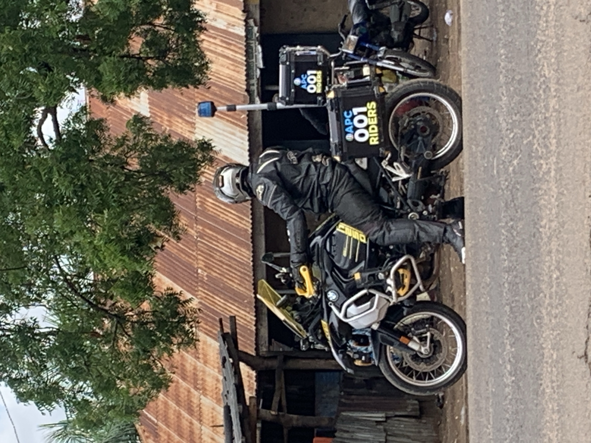 The image displays a black BMW R1200GS or R1250GS adventure motorcycle, with a rider in black motorcycle gear and a silver helmet, stationary on a paved road in Shendam, Nigeria. The motorcycle features yellow accents on the handguards and engine crash bars. Mounted on the rear are two black hard-shell panniers, each bearing the text "APC 001 RIDERS" in white and yellow lettering. A blue beacon light is affixed to a pole extending vertically from the rear of the bike. In the background, structures with heavily rusted corrugated iron roofs and weathered wooden framework are visible, partially obscured by dense green tree foliage. A second, smaller motorcycle is discernible further in the background to the right. The road surface in the foreground shows minor cracking.