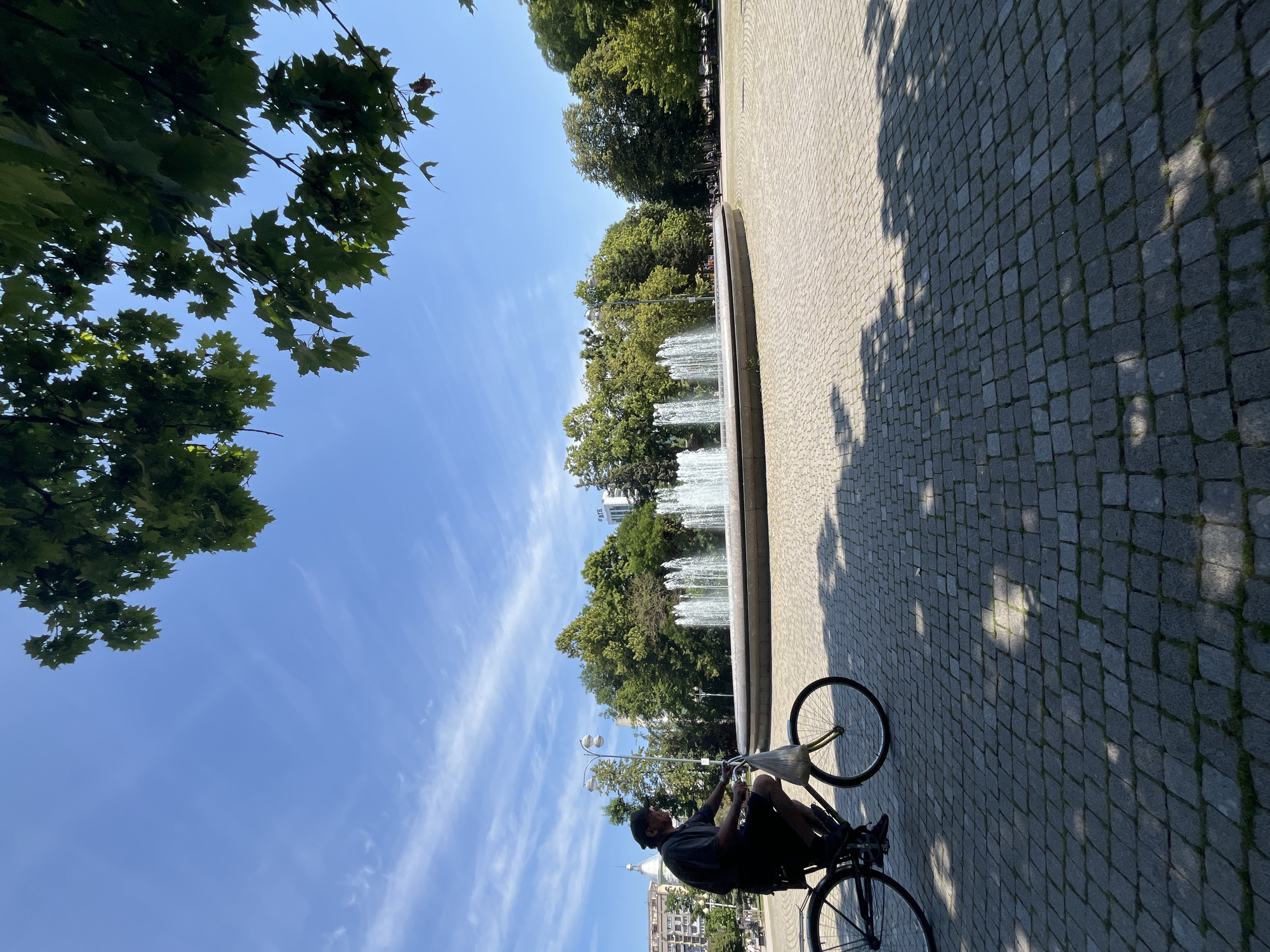 A man rides a bicycle on a cobblestone plaza in front of a large circular fountain with multiple streams of water shooting upwards. The fountain is surrounded by trees and a building with text that appears to be in Cyrillic script. The sky is blue with sparse white clouds, and leafy branches of trees overhang the scene from the top. The man is wearing dark clothing and a cap. The bicycle has a bag attached to the handlebars. The lighting suggests it is daytime, and shadows from trees are visible on the plaza. The specific location in Krasnodar, Russia cannot be determined from the visual information provided.