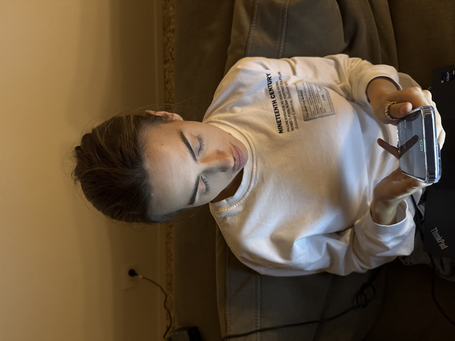 A female individual is seated on a light brown upholstered couch in an indoor, dimly lit environment with warm-toned lighting. Her light brown hair is styled up, and she wears a white long-sleeve shirt. The shirt displays text on the upper left chest, reading "NINETEENTH CENTURY", "PAULINE BORGHESE AS VENUS VICTRIX, SIGNED C. POSSI ROMA PORTRAIT SCULPTURE", "Antonio Canova, Italian art", and a graphic label "ITALIAN MYTH & MARBLE". She is looking down, holding a smartphone with a clear case in both hands. The phone's screen is illuminated, showing a charging icon, and casts light onto her face. A dark-colored Lenovo ThinkPad laptop is positioned below her hands. A ring is visible on the ring finger of her right hand. The background consists of a plain, light orange-brown wall. A black electrical outlet is visible on the wall, with a black power cable plugged into it, extending out of frame to the left. The specific location within Venice, Italy, cannot be determined from the image.
