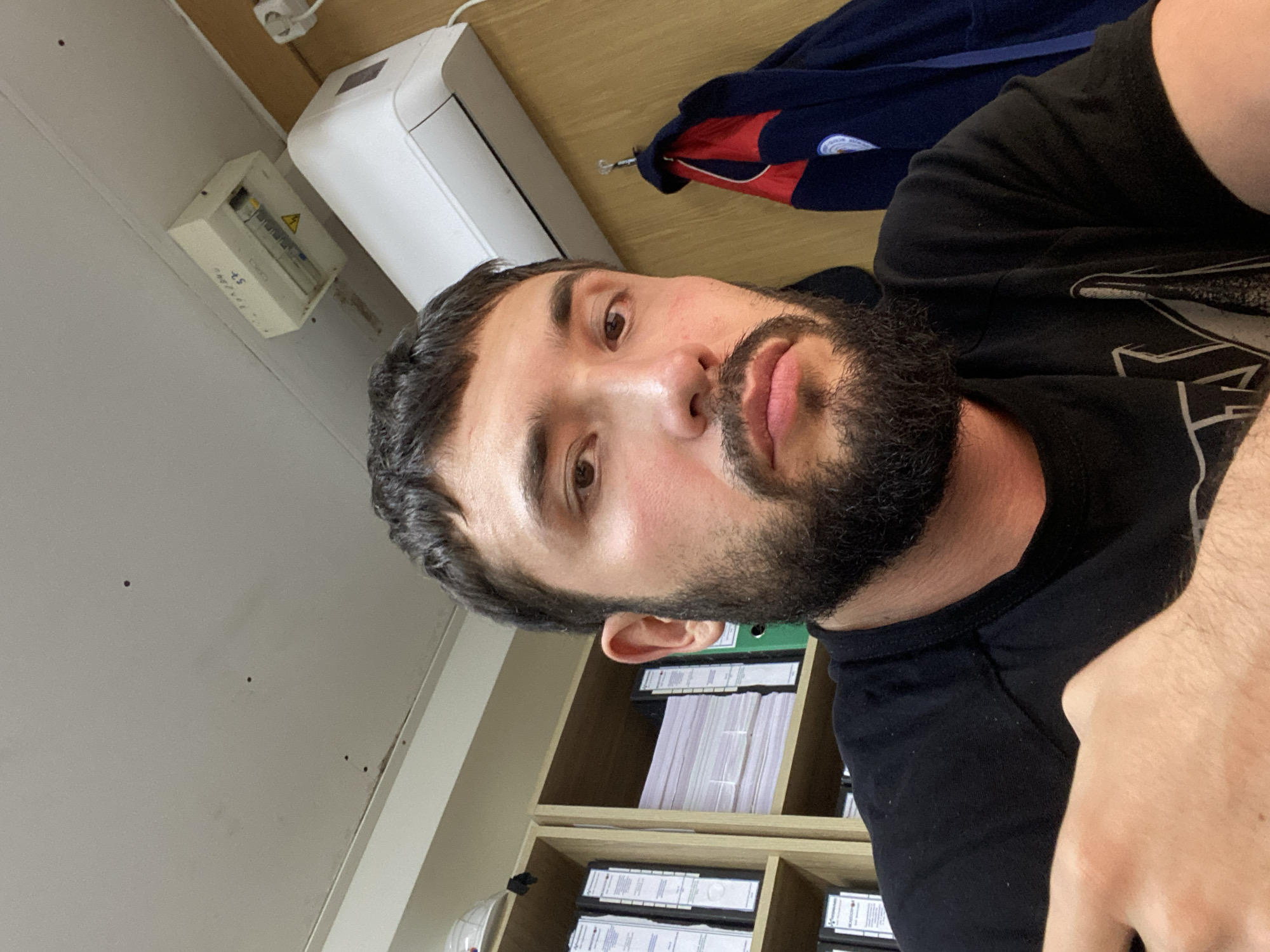 The image is a selfie taken by a man with a beard and short dark hair. He is wearing a black t-shirt and is sitting in an office environment. Behind him, there are shelves filled with binders and folders. An air conditioning unit is mounted on the wall above him. A jacket is hanging on a hook to the right of the frame. The ceiling shows some small dark spots. There is a white electrical box mounted on the ceiling, with some writing on it. The overall lighting suggests an indoor setting. The location is identified as Luzhitsy, Russia.