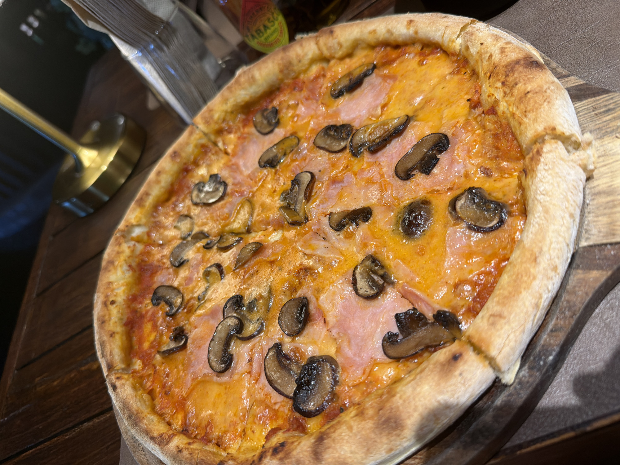 A high-angle, medium shot depicts a mushroom and ham pizza on a wooden board. The pizza has a thick, golden-brown crust and is topped with tomato sauce, cheese, ham, and sliced mushrooms. It is sliced into approximately six pieces. The pizza is resting on a dark wooden board. In the background, out of focus, are a stack of napkins, a golden-colored metallic object, and a bottle of Tabasco sauce. The scene appears to be indoors. The image was taken in Baan Chaweng, Thailand.